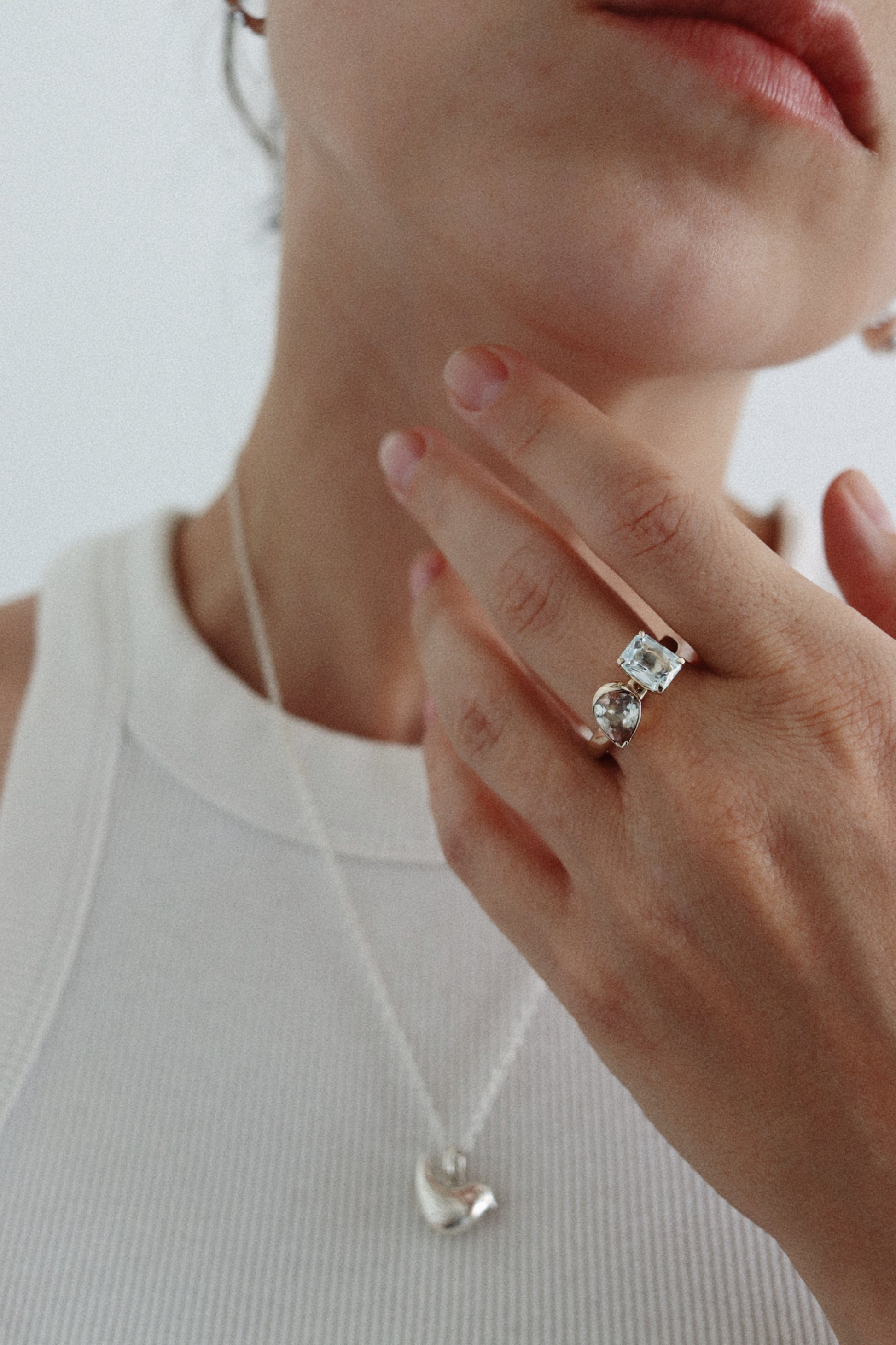 A close-up lifestyle shot of a model wearing a silver ring with a square-cut clear gemstone on her finger, paired with a silver heart pendant necklace and a white ribbed tank top.