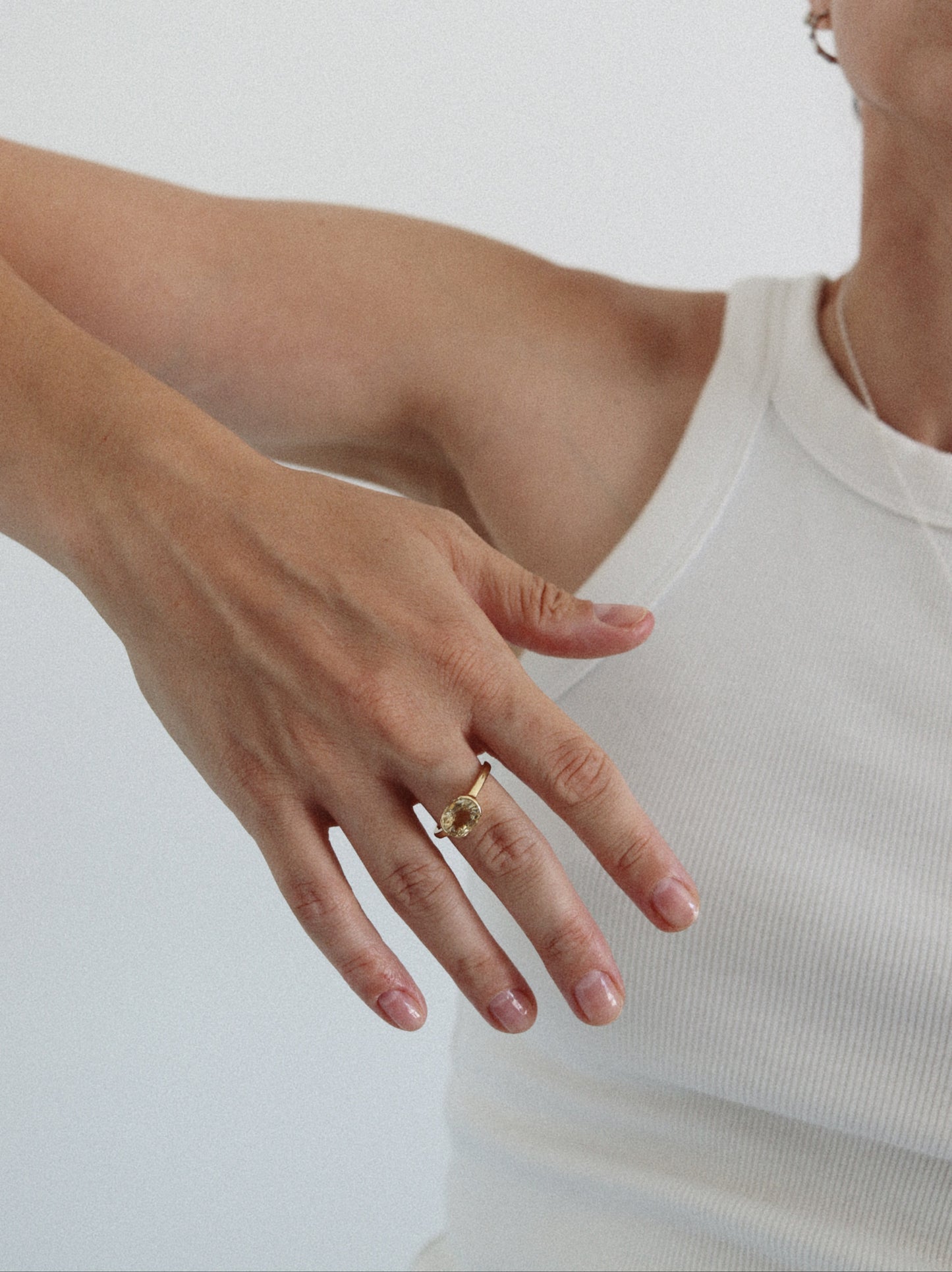 A close-up of a person's hand wearing a delicate gold ring with small clear stones, paired with a white ribbed tank top against a plain light background.