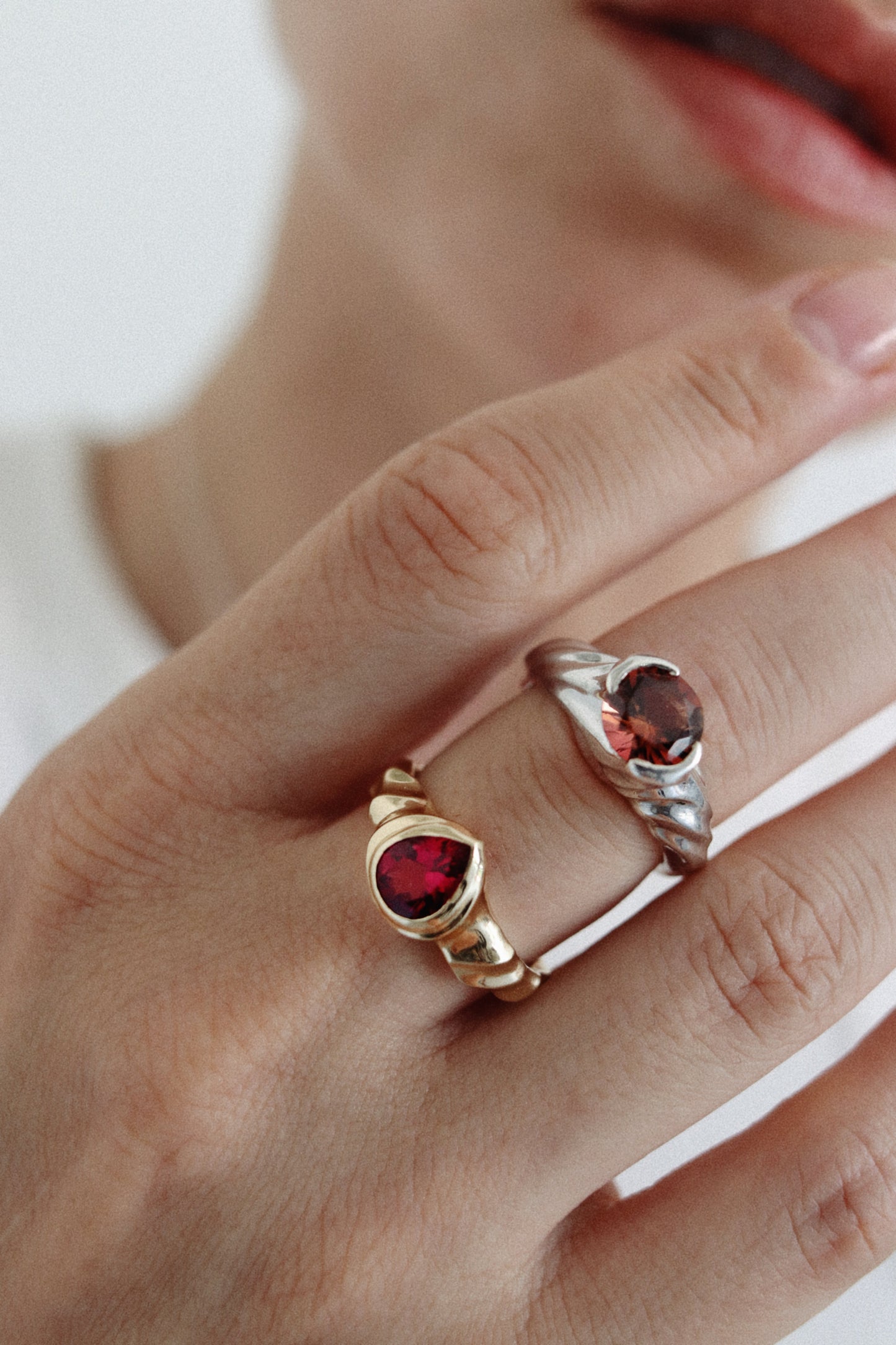 A close-up of a person's hand wearing two rings: one gold ring with a teardrop-shaped red gemstone and one silver ring with a round red gemstone.