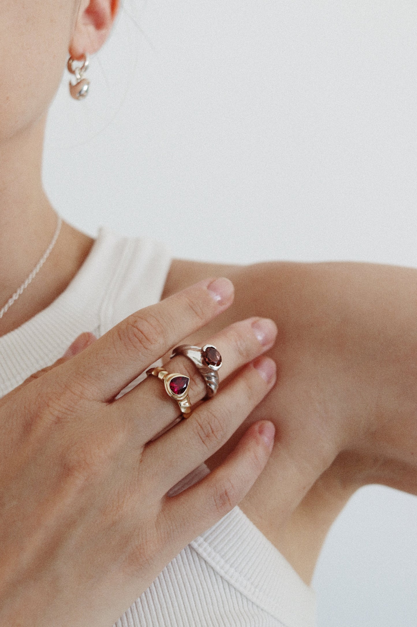 A close-up lifestyle shot of a model wearing two gold rings with heart-shaped red gemstones on her fingers, paired with a white ribbed top against a clean white background.