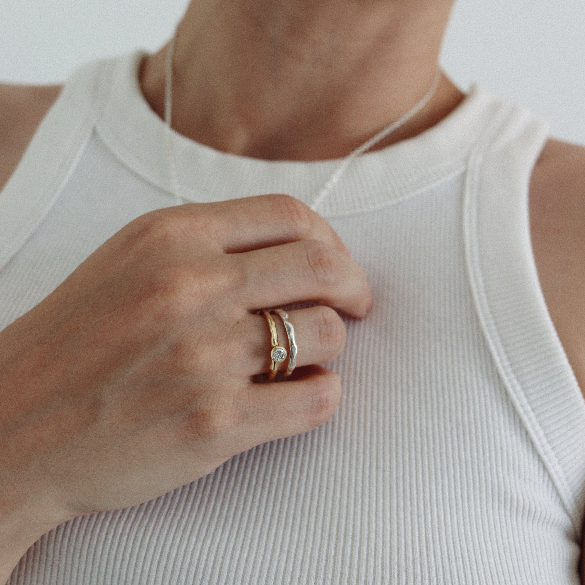 A close-up of a person wearing a delicate gold ring with a small clear gemstone on their finger, paired with a white ribbed tank top and a thin silver necklace.