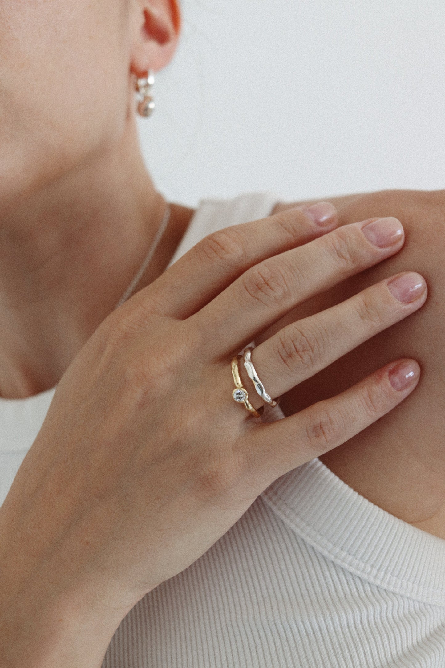 A close-up of a person's hand resting on their shoulder, wearing a delicate gold ring with a small clear gemstone. The person is wearing a white ribbed top, and a small gold hoop earring is visible on their ear.