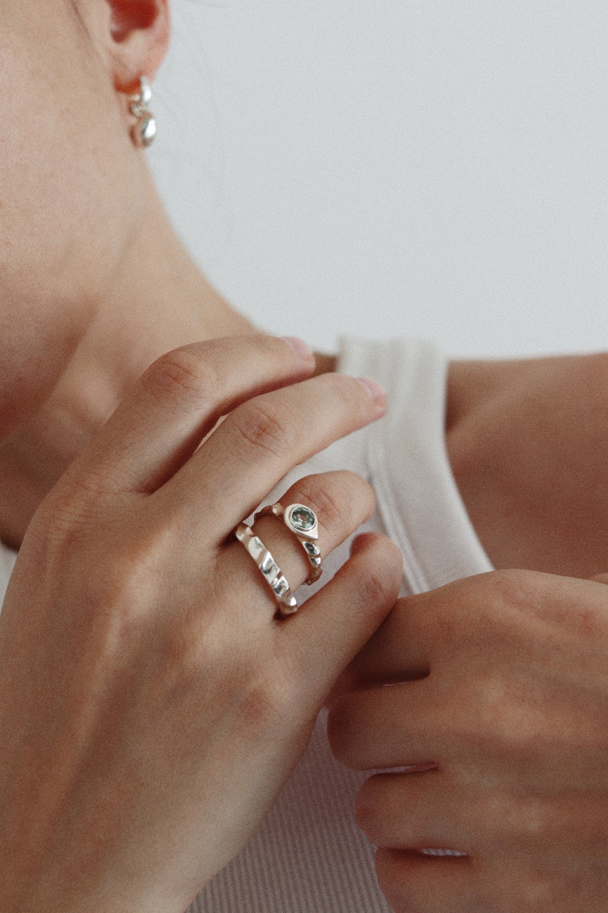 A close-up lifestyle shot of a person wearing a silver ring with a teardrop-shaped clear gemstone and a textured silver band, paired with a matching textured silver hoop earring.