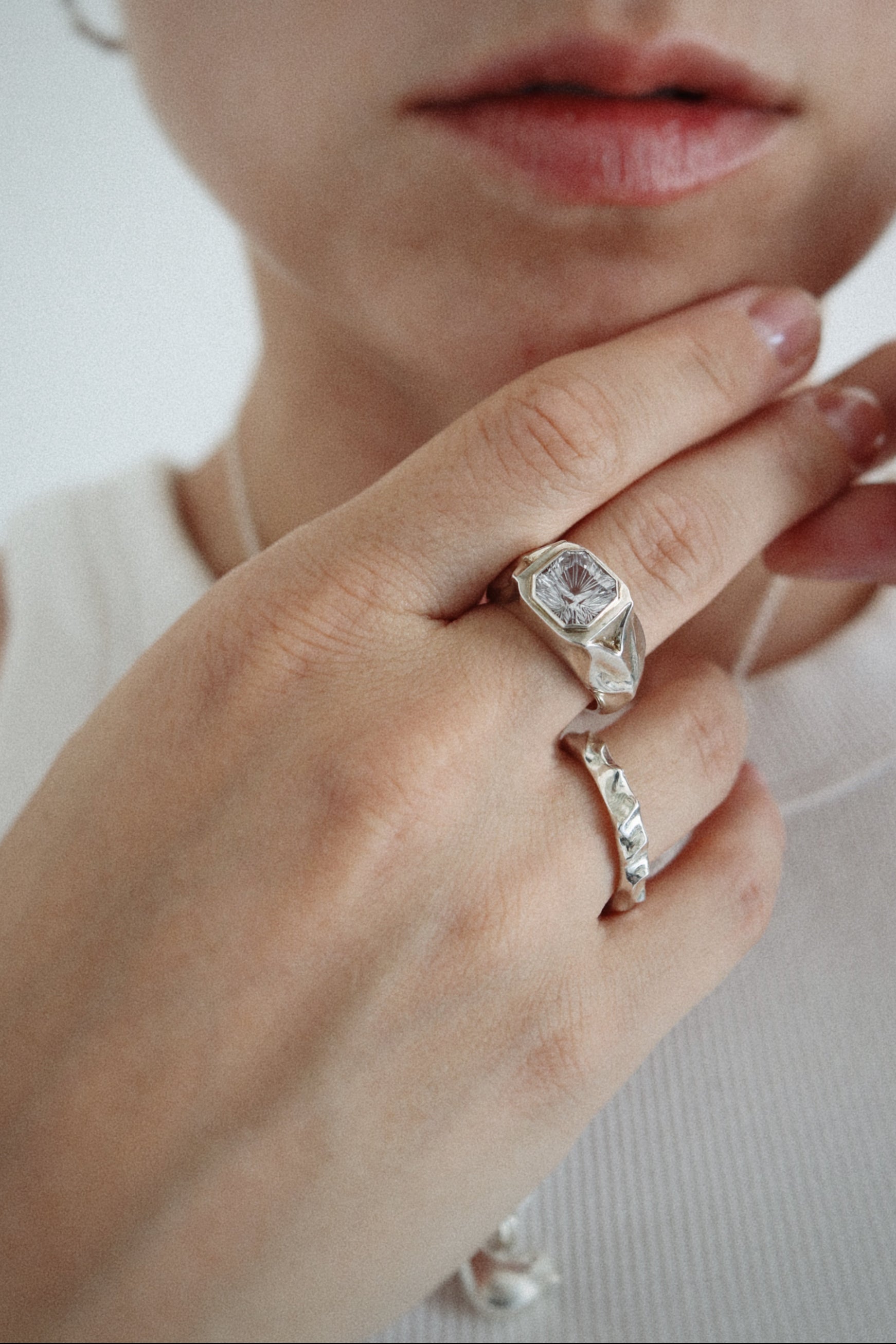 A close-up lifestyle shot of a person wearing a silver signet ring with a clear rectangular gemstone on their middle finger, with another silver ring on their ring finger, set against a clean white background.