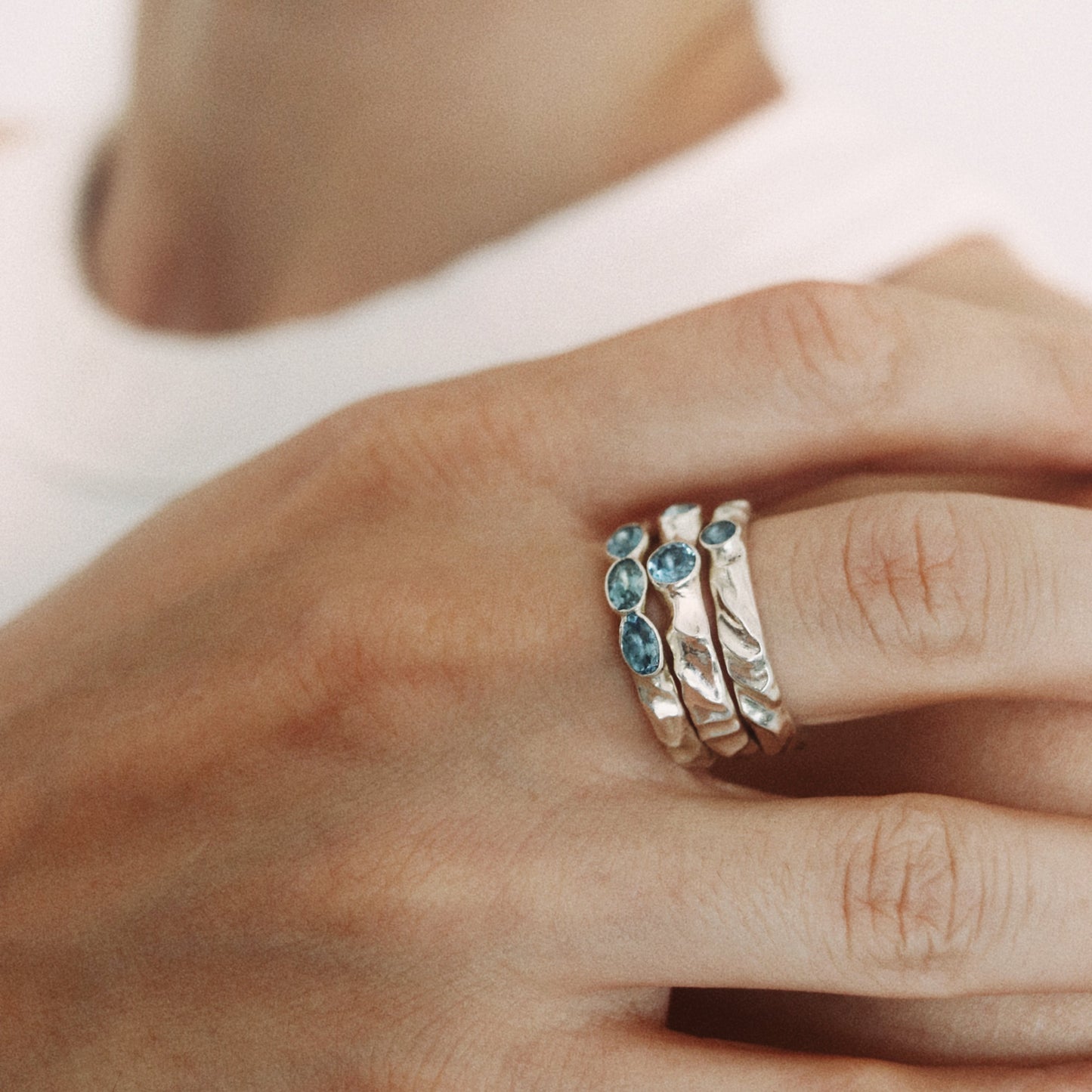 A close-up of a person's hand wearing three stacked silver rings, each accented with small round blue gemstones, against a soft white background.