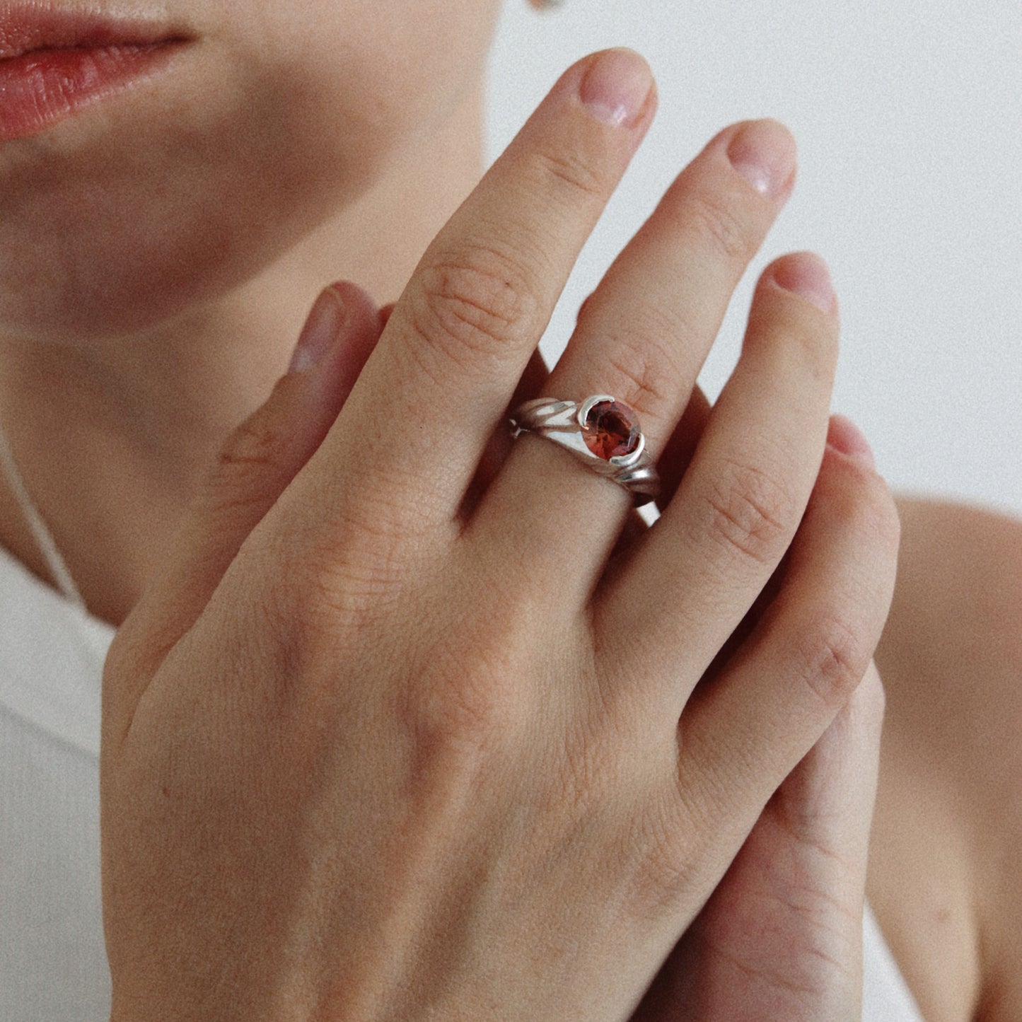 A close-up of a person's hand wearing a silver ring with a small oval pink gemstone, against a soft light gray background.