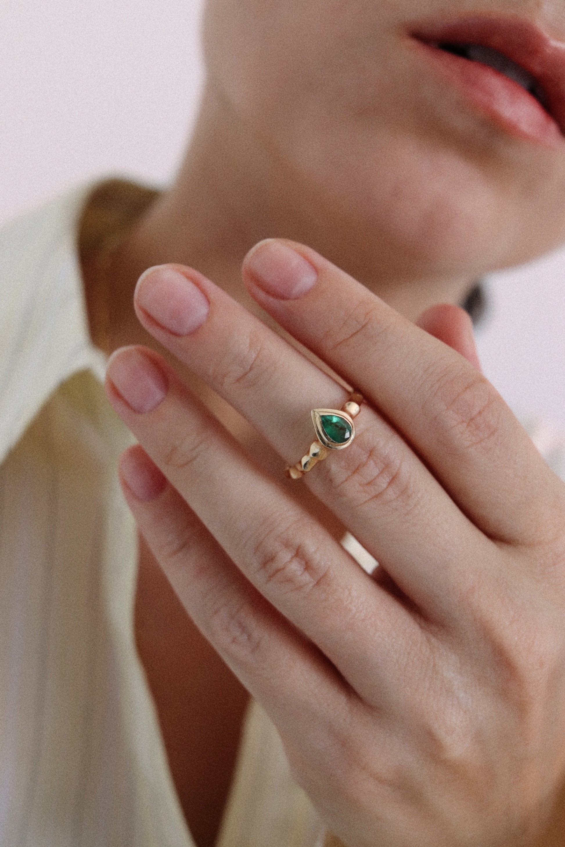 A close-up of a person's hand wearing a gold ring with a teardrop-shaped green gemstone, set against a soft-focus background of a person's face and a cream-colored blouse.