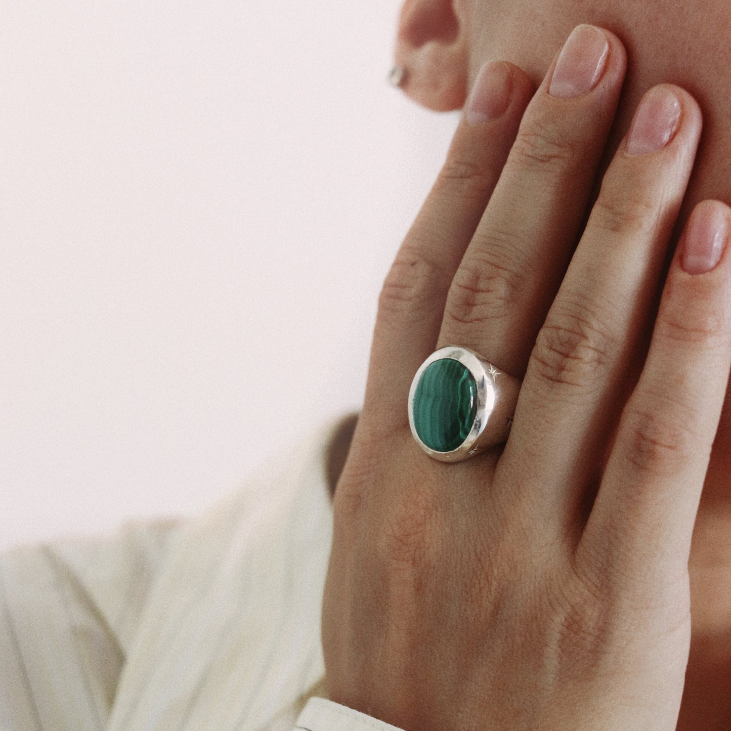 A close-up of a person's hand wearing a silver ring with an oval green malachite stone, set against a soft, light background.