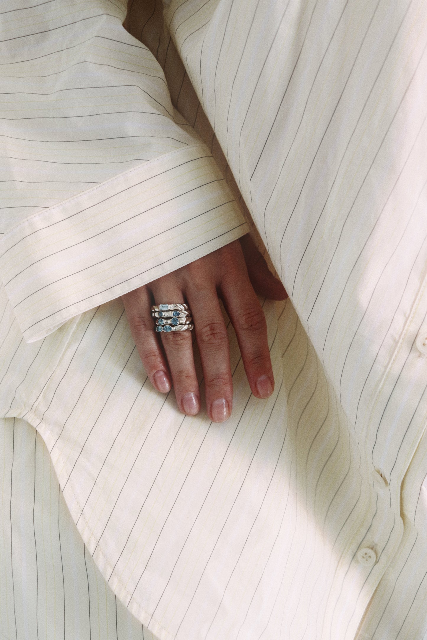 A close-up of a hand wearing a silver ring with clear stones, resting on a cream-colored pinstriped shirt.