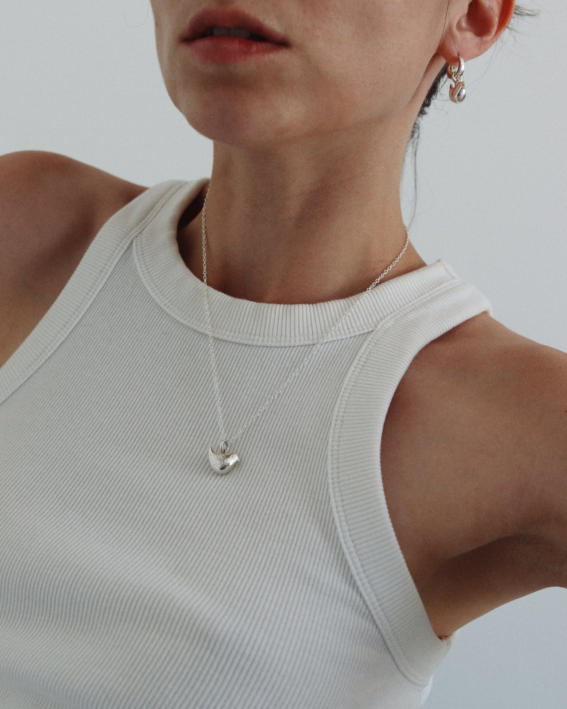 A close-up of a model wearing a delicate silver necklace with a small heart-shaped pendant and a matching silver hoop earring, paired with a white ribbed tank top against a light gray background.