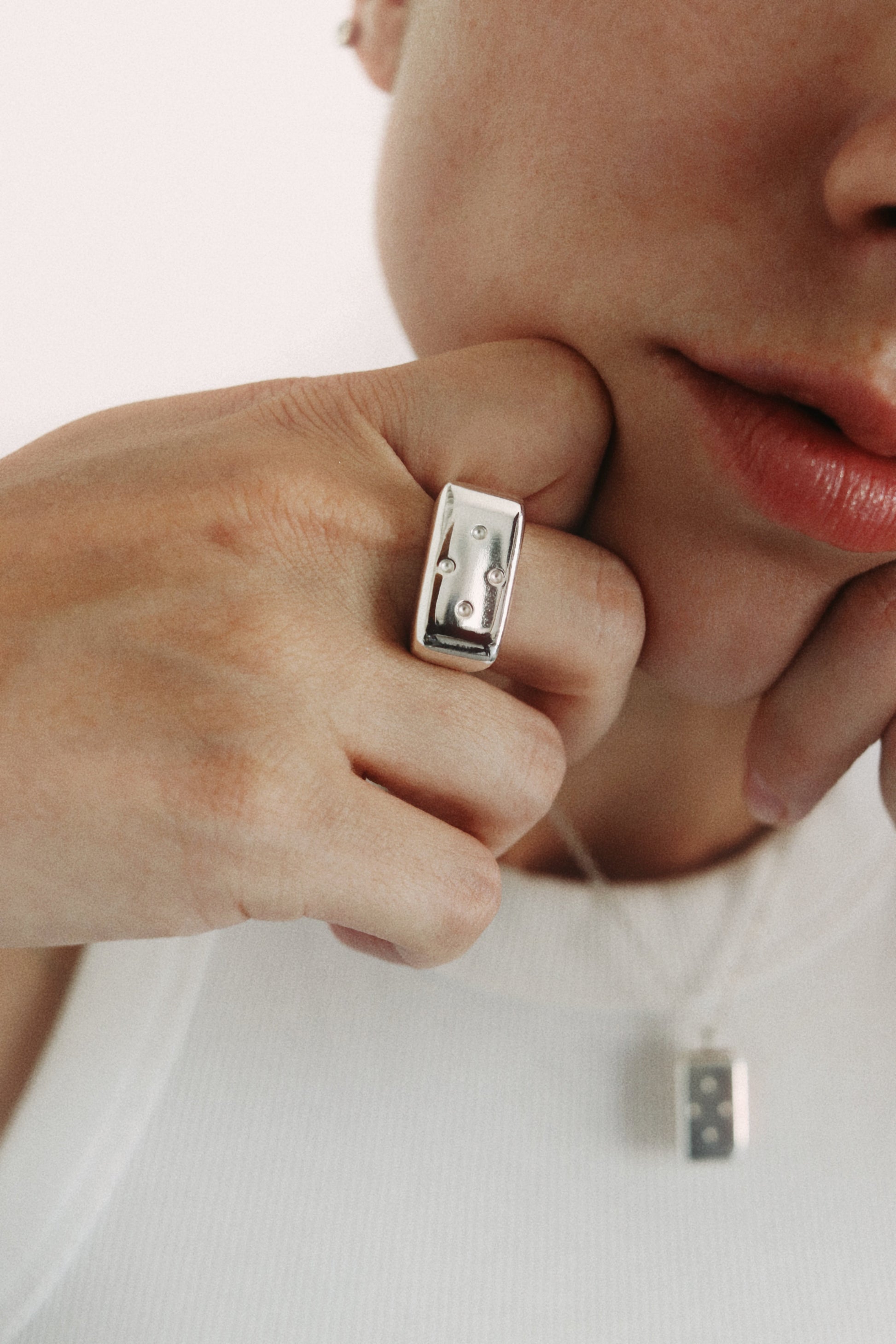 A close-up lifestyle shot of a person wearing a chunky silver rectangular ring on their finger, with a matching silver pendant necklace visible against a white ribbed top.