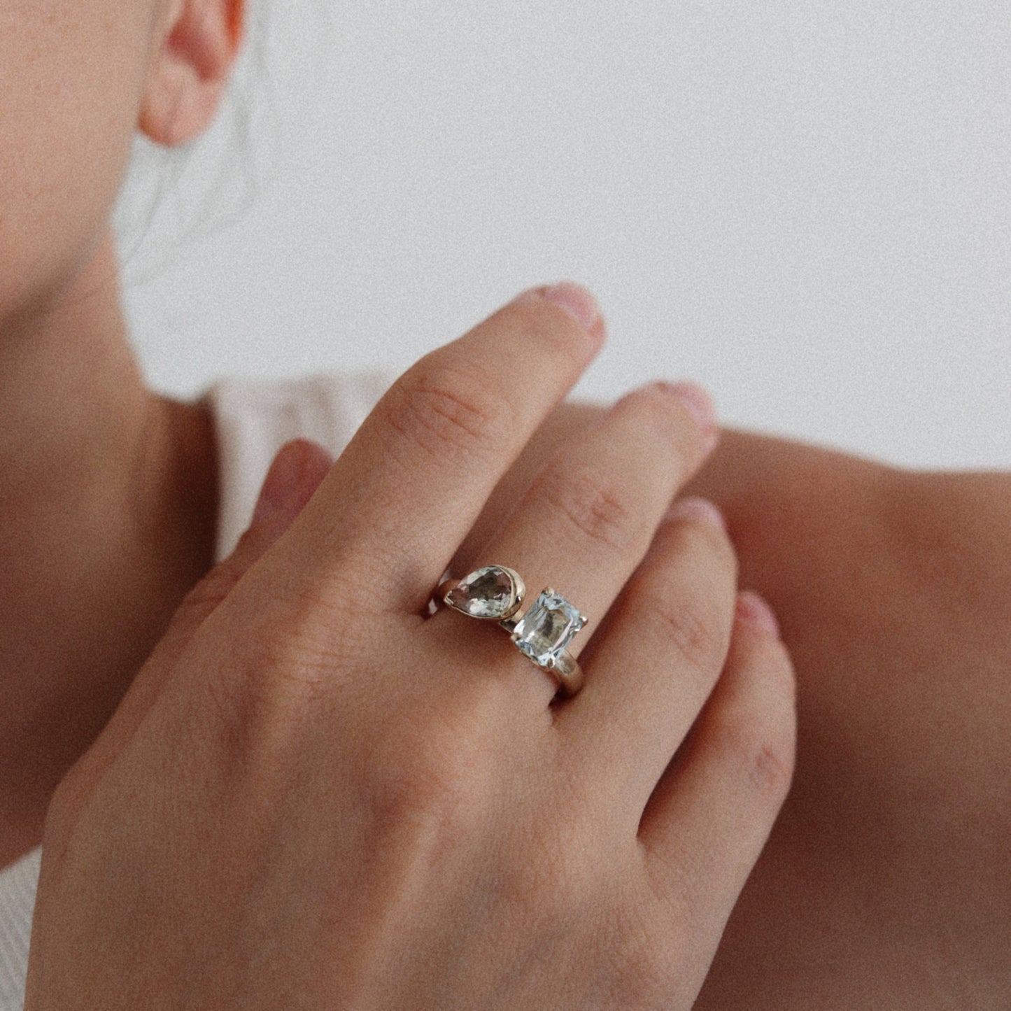 A close-up of a person's hand wearing a silver ring with two clear gemstones, one pear-cut and one rectangular-cut, against a soft light gray background.