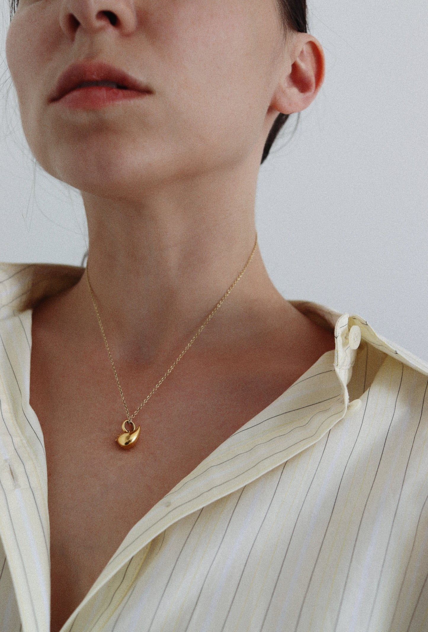 A close-up of a model wearing a delicate gold chain necklace with a small gold leaf pendant, styled with a cream-colored striped button-down shirt.