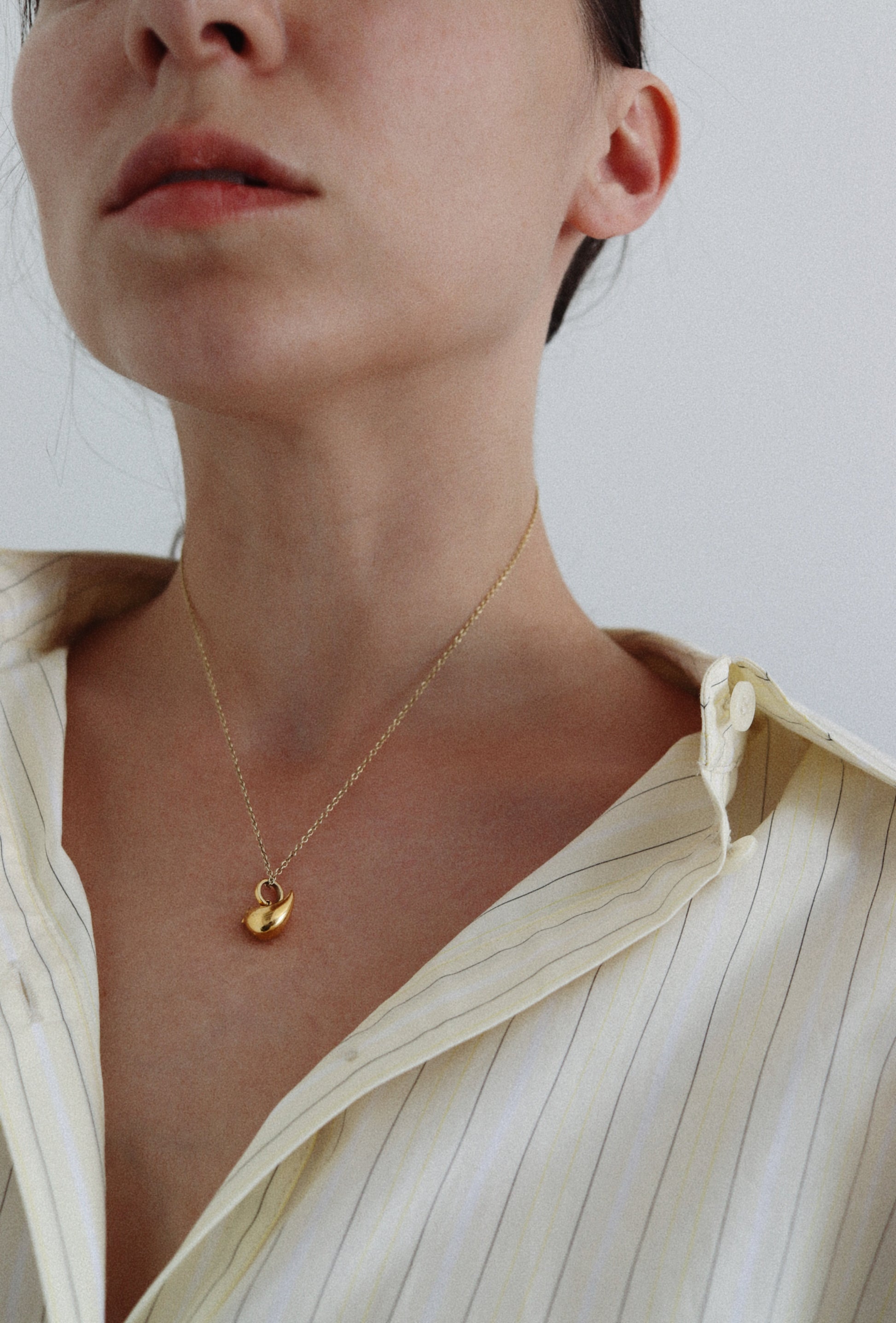 A close-up of a model wearing a delicate gold chain necklace with a small gold leaf pendant, styled with a cream-colored striped button-down shirt.