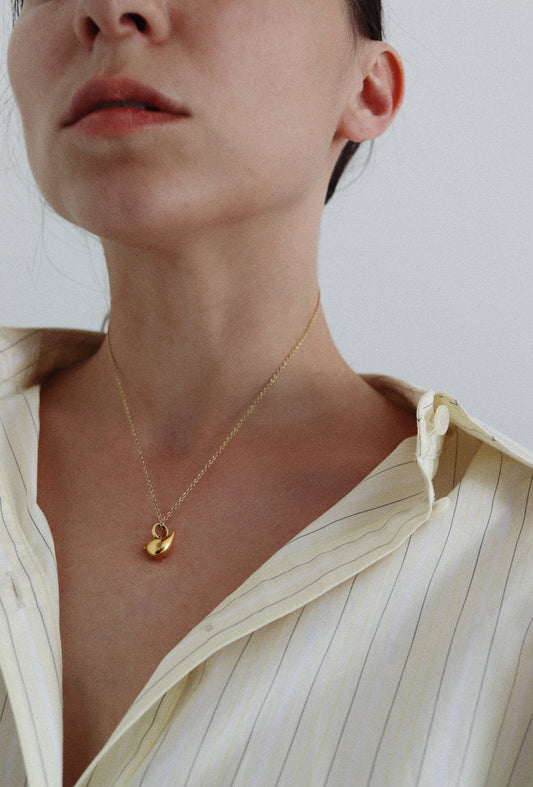 A close-up of a model wearing a delicate gold chain necklace with a small gold leaf pendant, styled with a cream-colored striped button-down shirt.