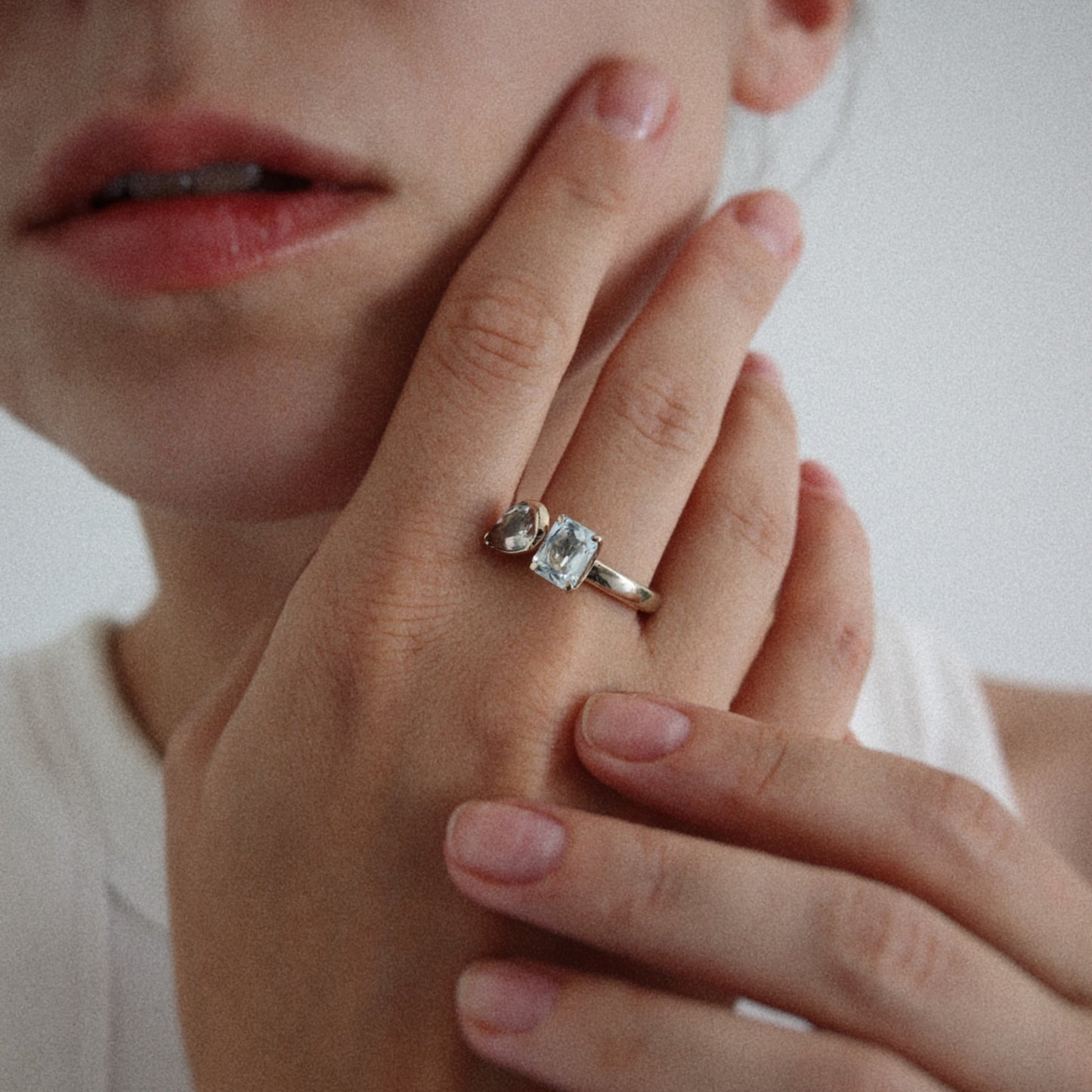A close-up lifestyle shot of a person wearing a silver ring with a cushion-cut clear gemstone on their finger, with their hand resting near their face against a plain light background.