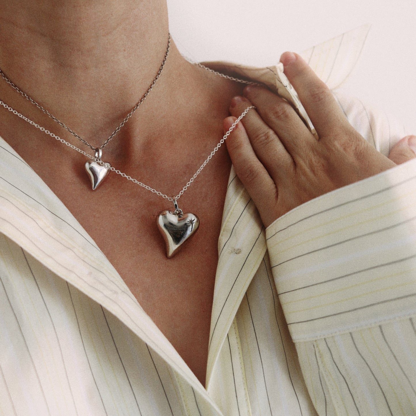 A close-up of a person wearing two silver heart-shaped pendant necklaces over a cream-colored pinstriped shirt.