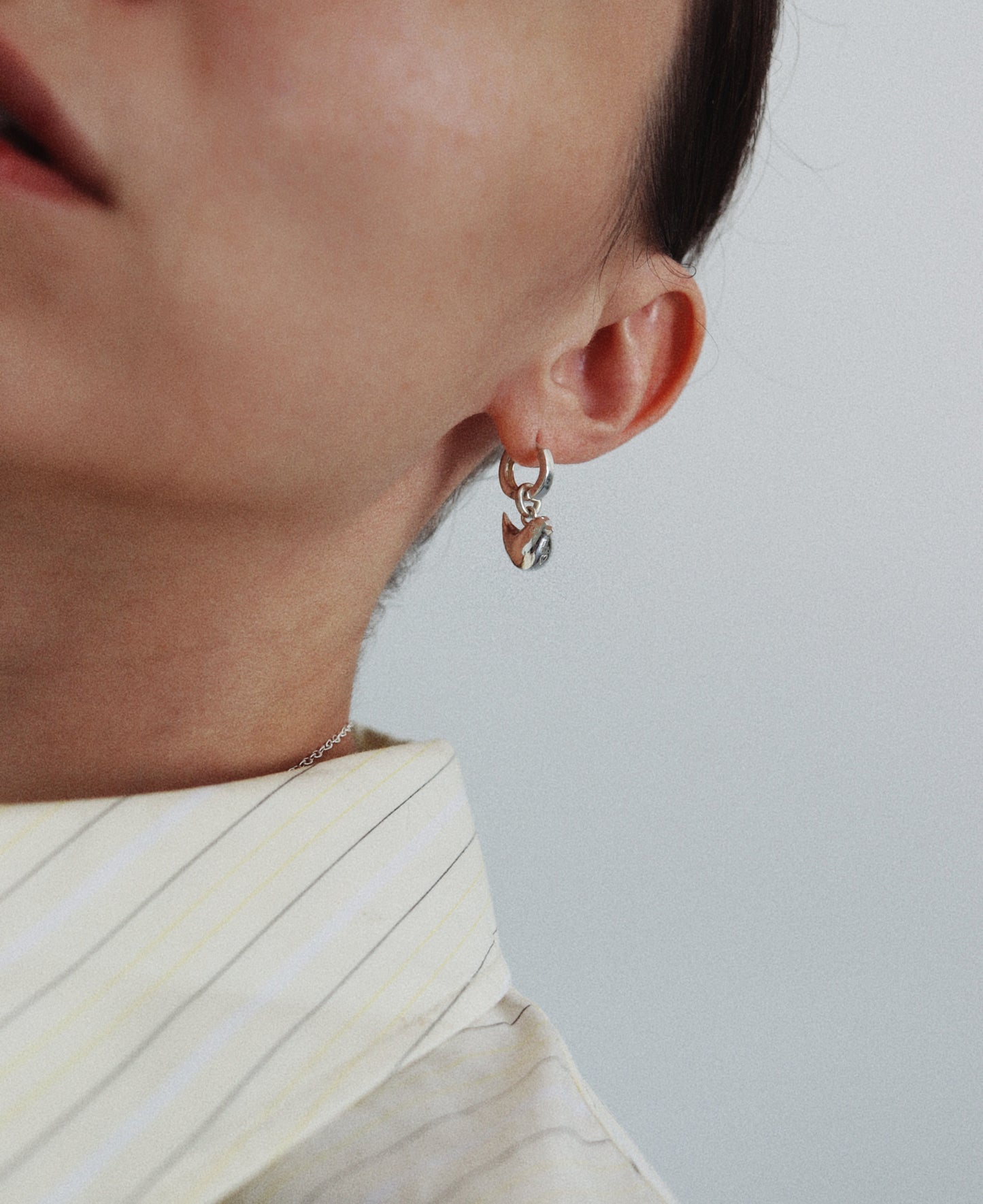 A close-up of a person wearing a small silver hoop earring with a dangling heart-shaped charm, paired with a cream-colored pinstriped shirt against a light gray background.