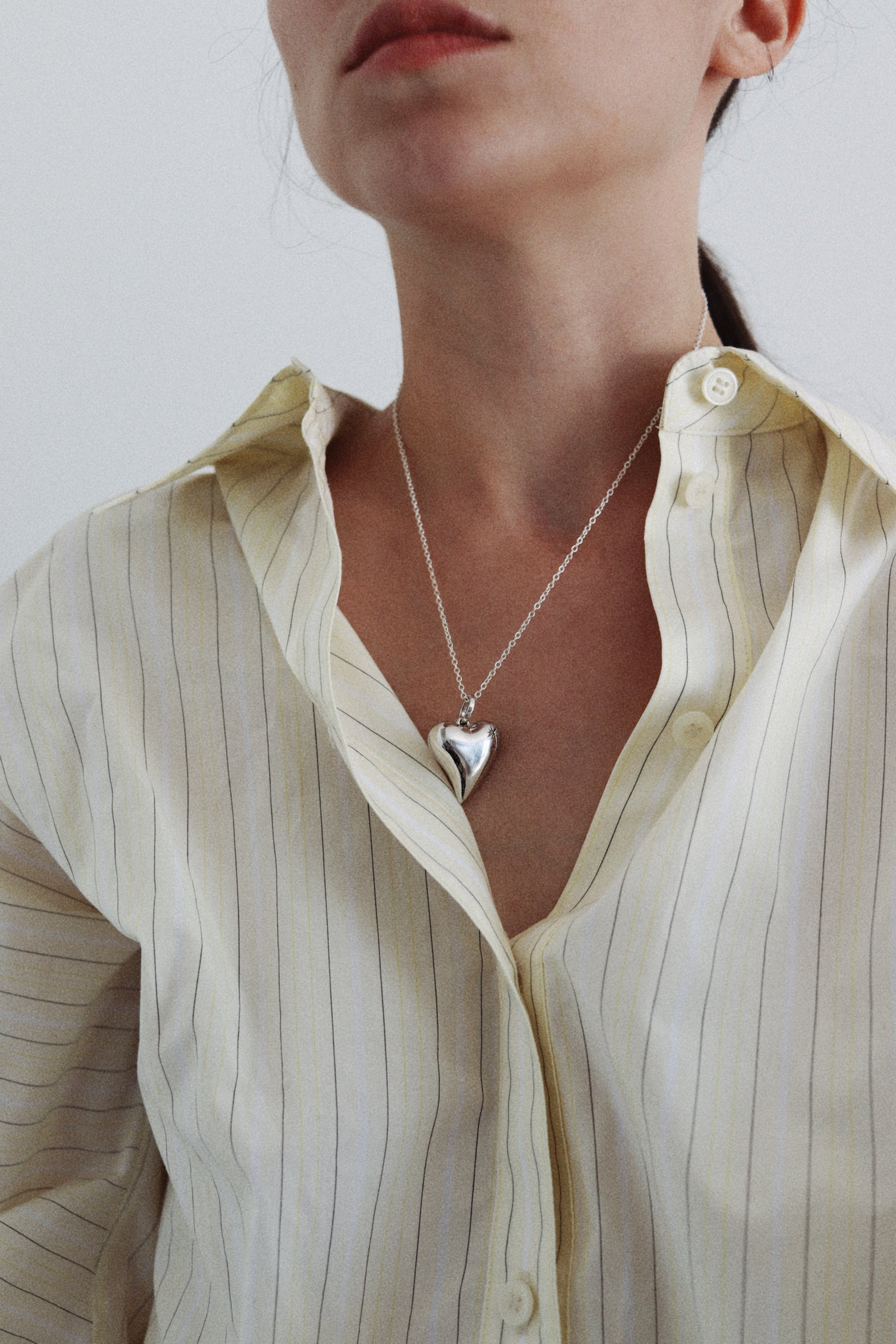 A close-up of a model wearing a silver heart pendant necklace over a cream-colored, vertically striped button-down shirt.