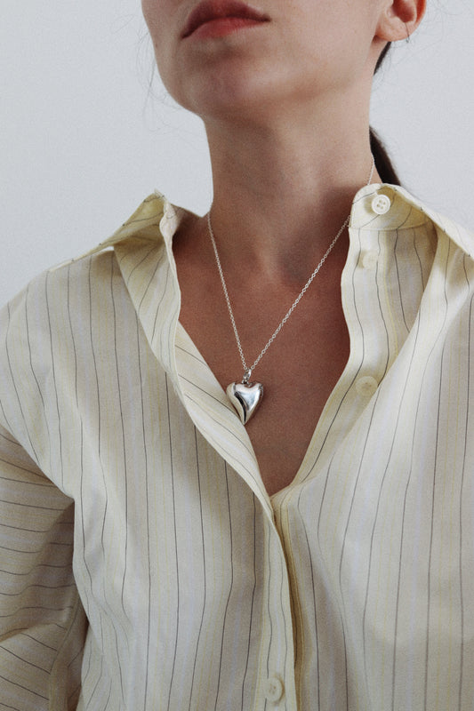 A close-up of a model wearing a silver heart pendant necklace over a cream-colored, vertically striped button-down shirt.