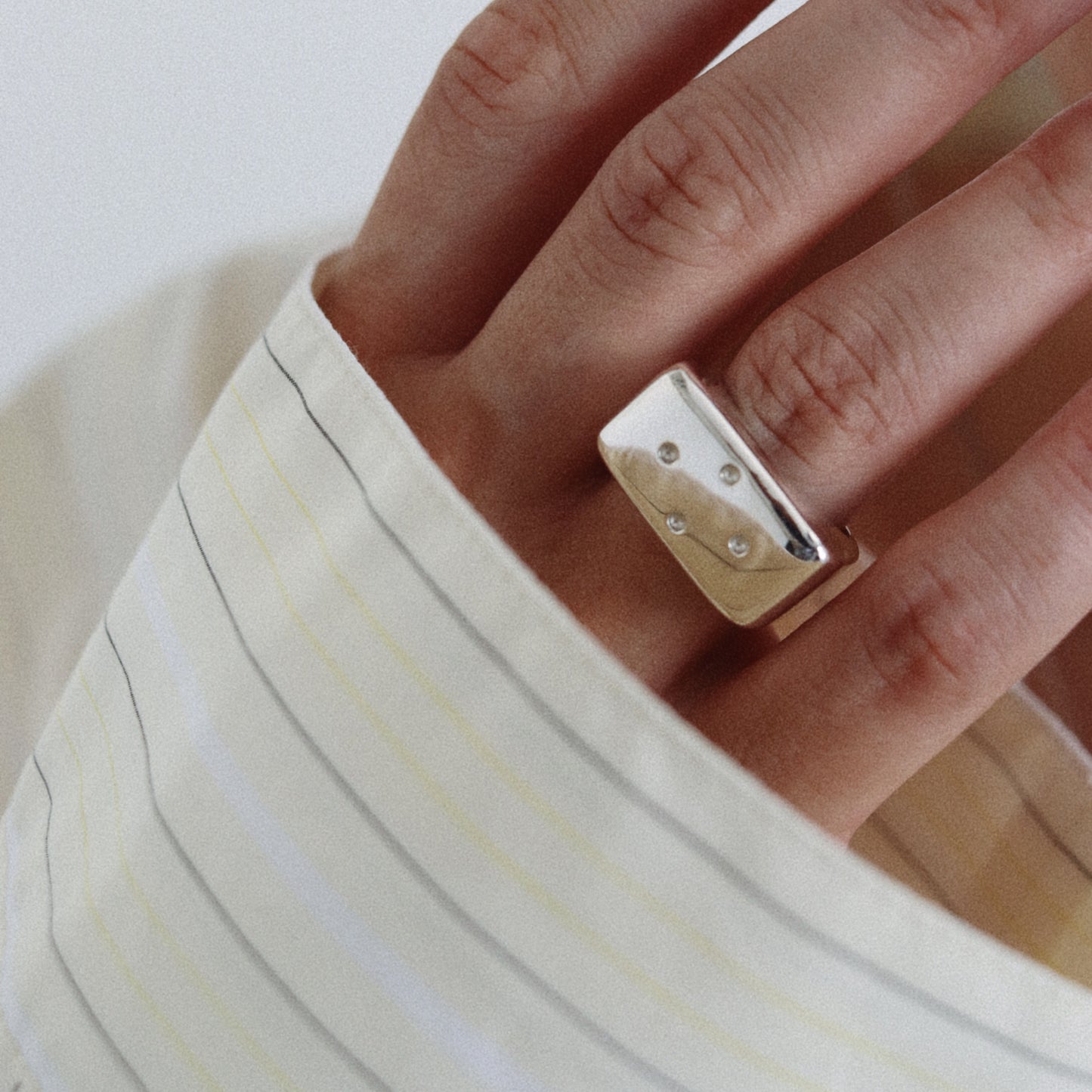 A close-up of a person's hand wearing a chunky, polished gold ring with small clear stones, styled with a cream-colored striped shirt.