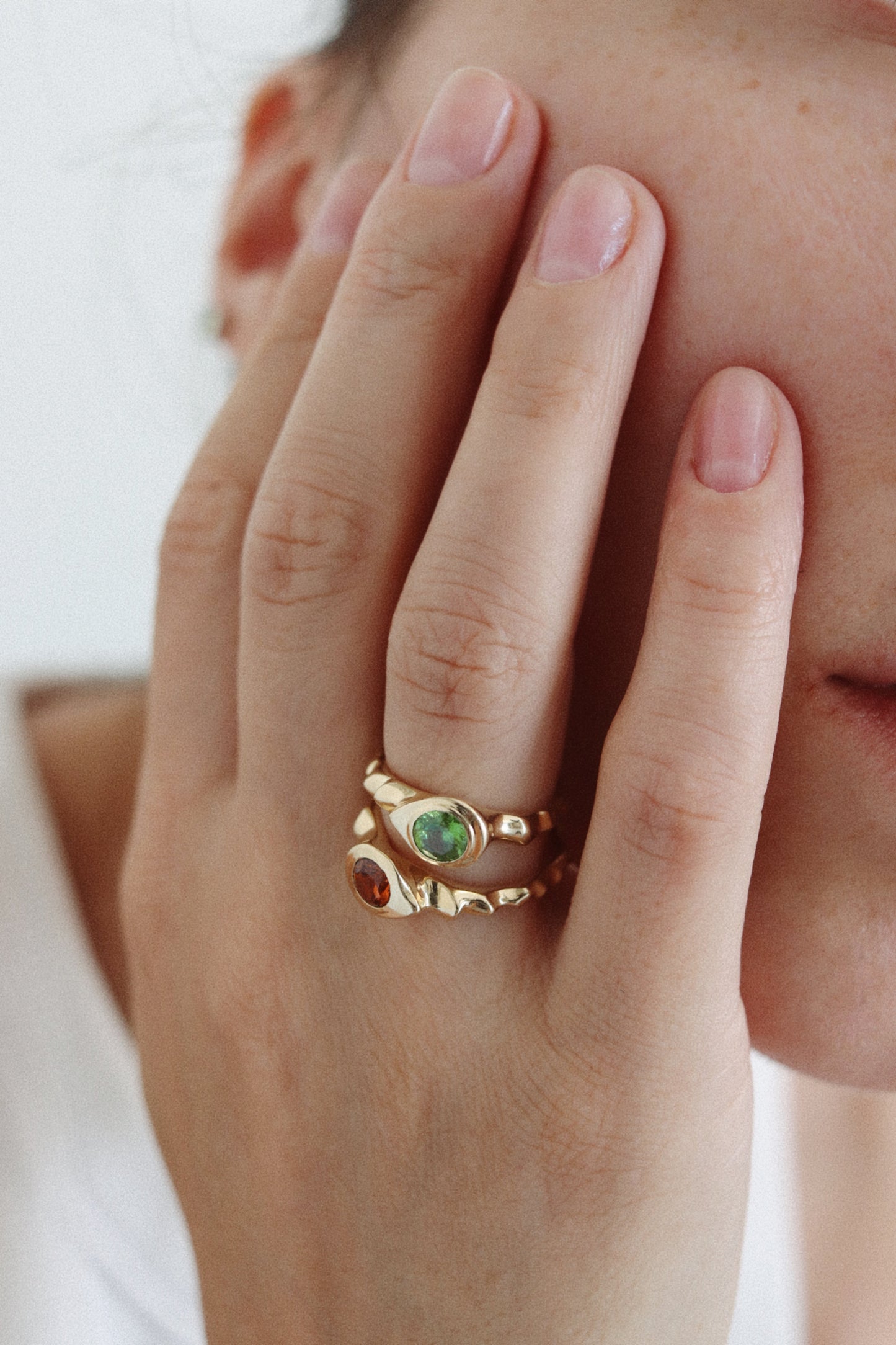 A close-up lifestyle shot of a person's hand wearing a gold ring with a green oval stone and a smaller red oval stone.