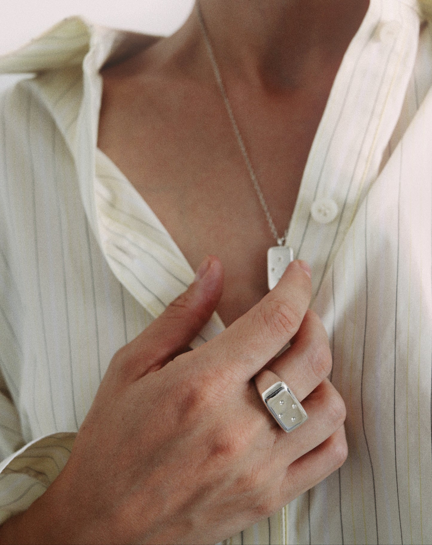 A close-up lifestyle shot of a person wearing a silver rectangular pendant necklace and a matching silver ring on their finger, styled with a cream-colored striped button-up shirt.