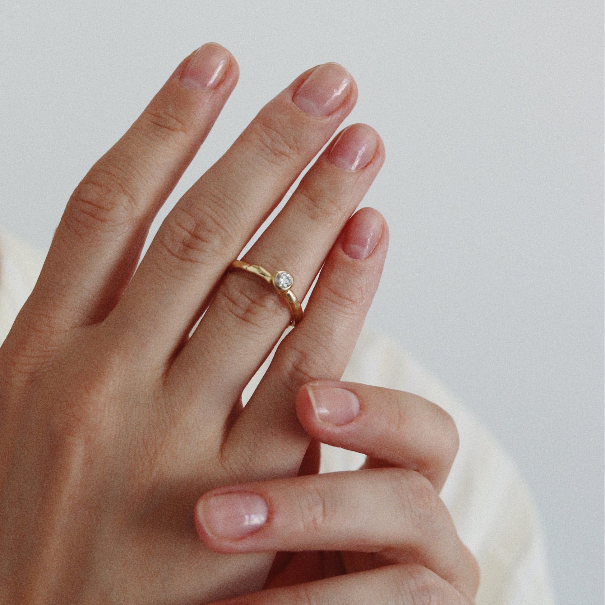 A close-up of a person's hands wearing a delicate gold ring with a small clear gemstone on the ring finger, set against a soft light gray background.