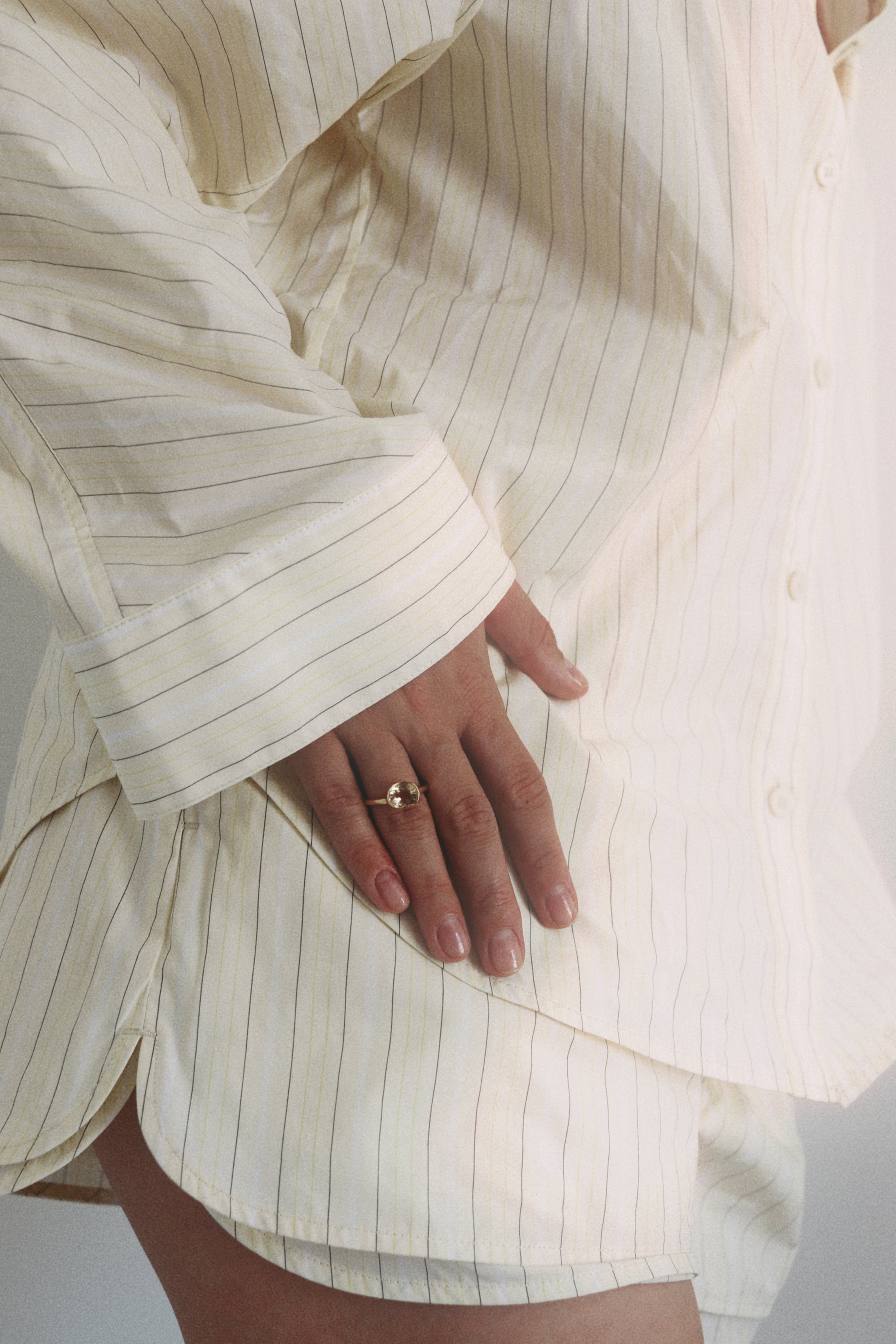 Close-up of a person wearing a cream-colored pinstriped button-up shirt and matching shorts, with a gold ring on their finger.