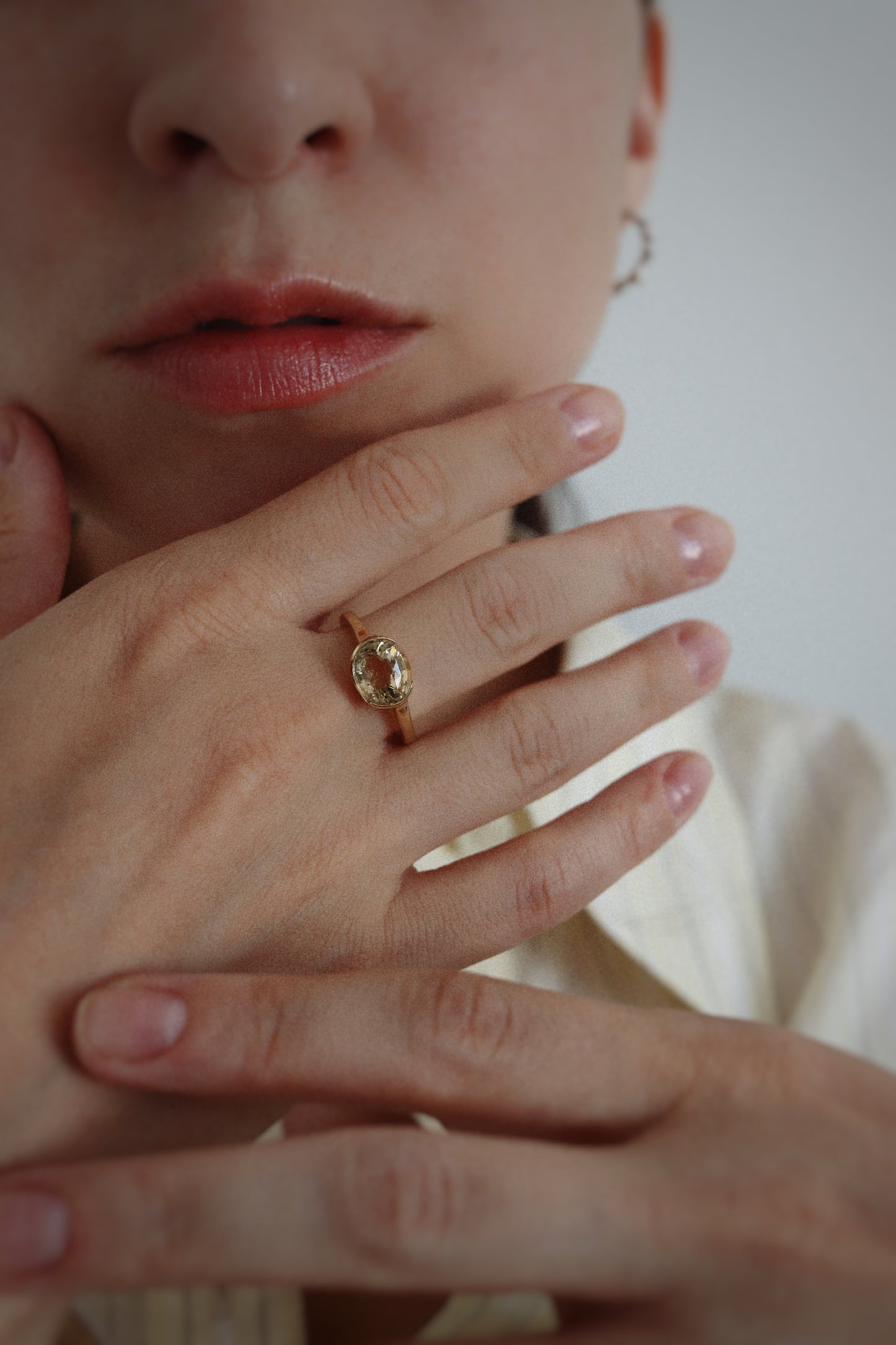 A close-up lifestyle shot of a person wearing a gold ring with a textured, organic-shaped setting and small clear stones, resting their hand on their chin.