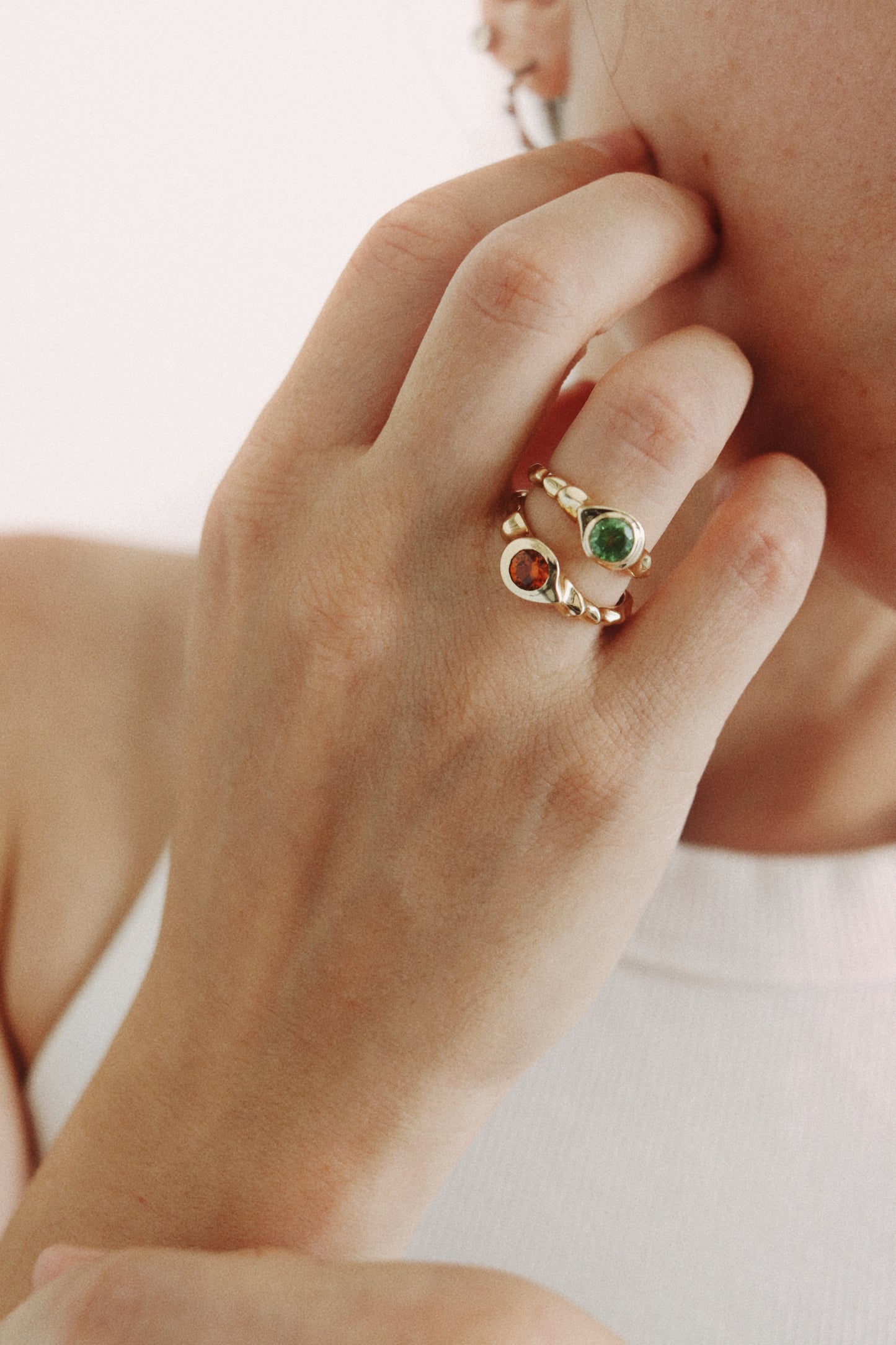 A close-up lifestyle shot of a person's hand wearing a gold ring with a double circular design, one featuring a green stone and the other an orange stone.