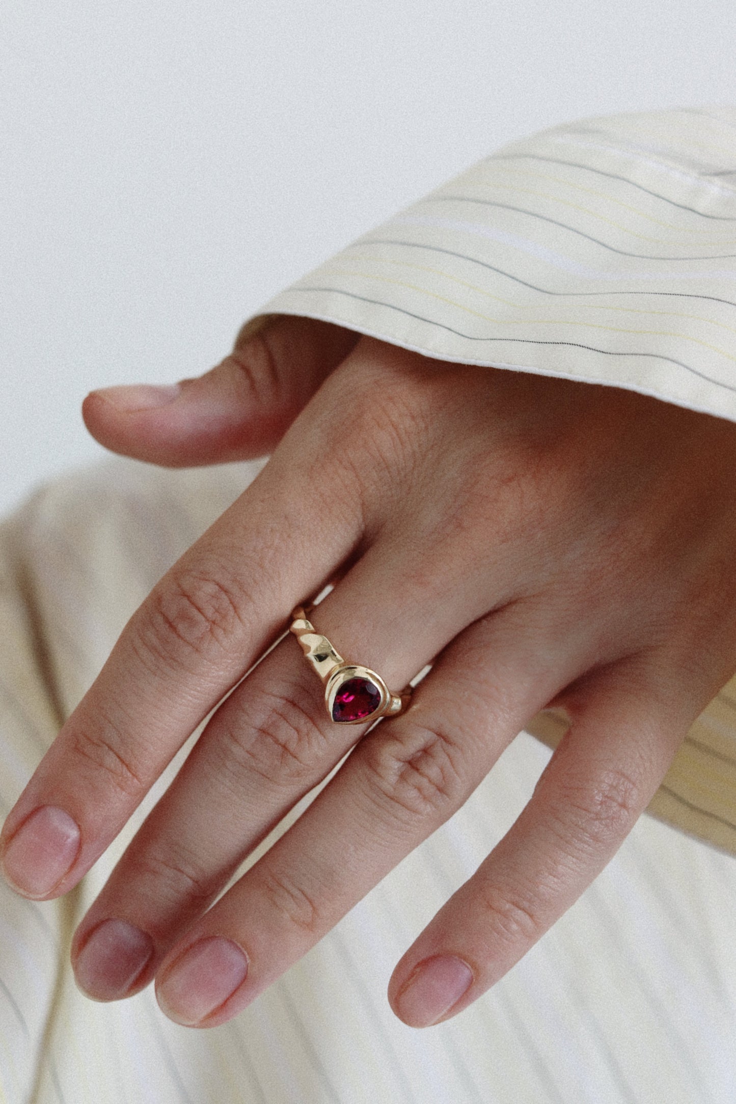 A gold ring with a heart-shaped red gemstone worn on a person's ring finger, shown against a soft cream-colored striped shirt and light background.