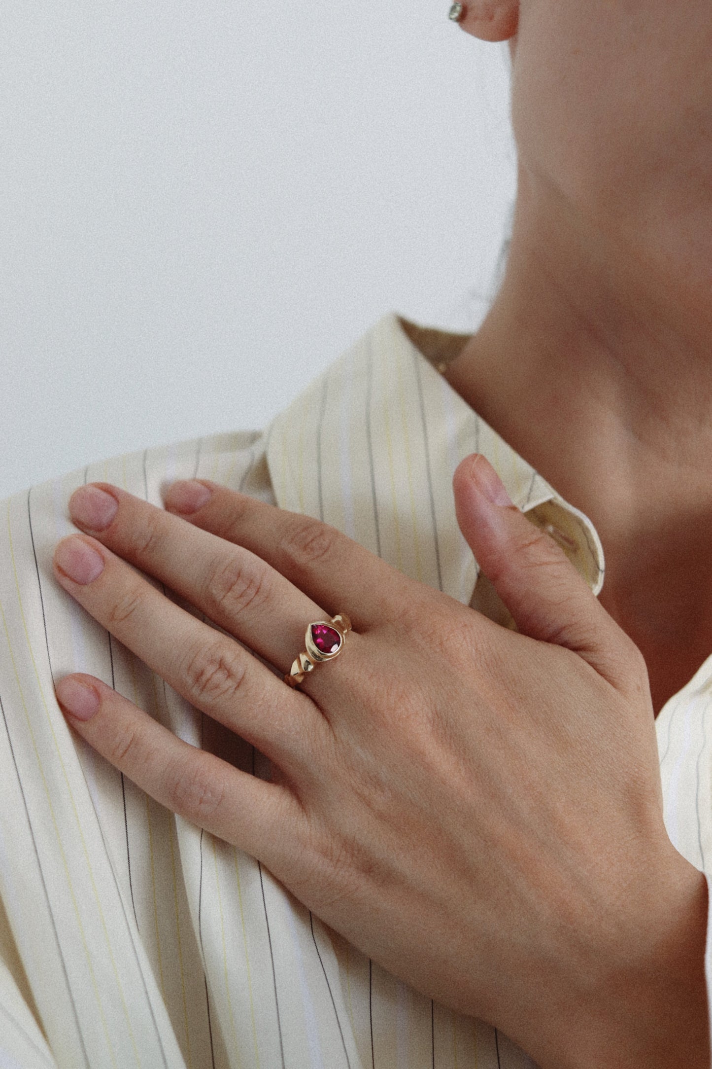A close-up lifestyle shot of a person wearing a gold ring with a marquise-cut red gemstone on their hand, resting on their striped cream shirt.