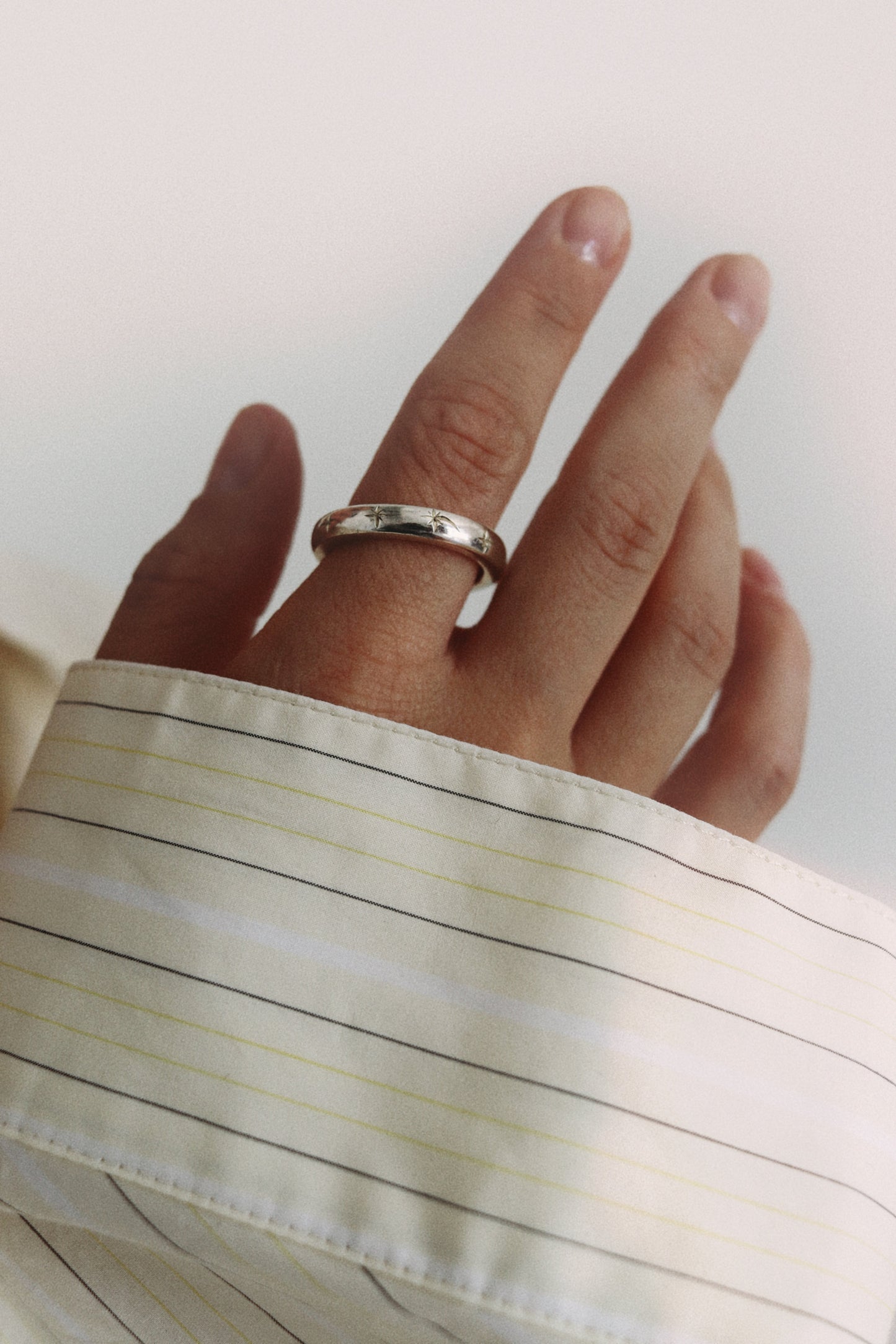 A silver band ring with small engraved letters, worn on a person's ring finger. The person is wearing a cream-colored dress shirt with thin dark stripes.
