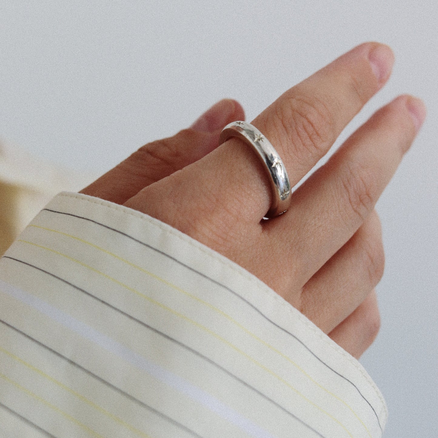 A close-up of a hand wearing a simple polished silver band ring, with a cream-colored striped shirt cuff visible.