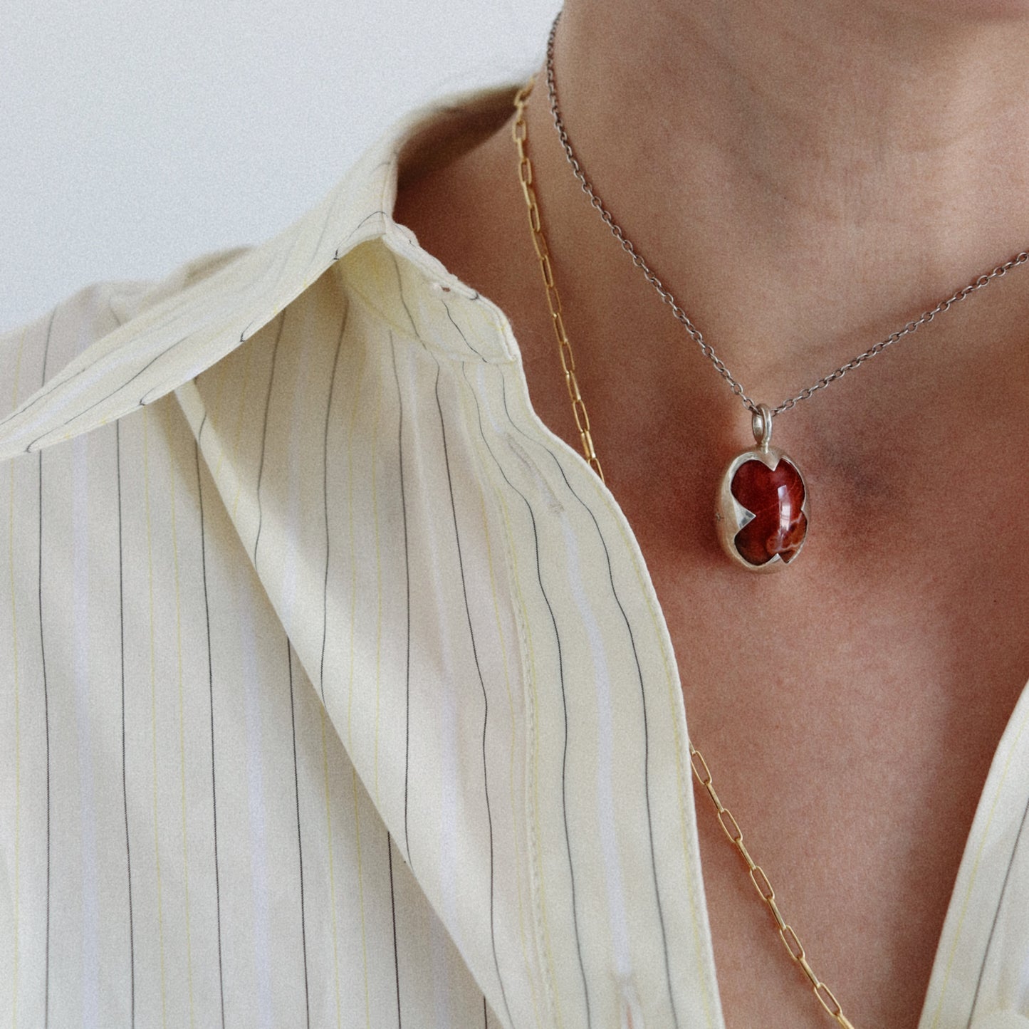 A close-up of a person wearing a silver-toned necklace with a red clover-shaped pendant, layered over a gold chain, paired with a cream-colored pinstriped shirt.