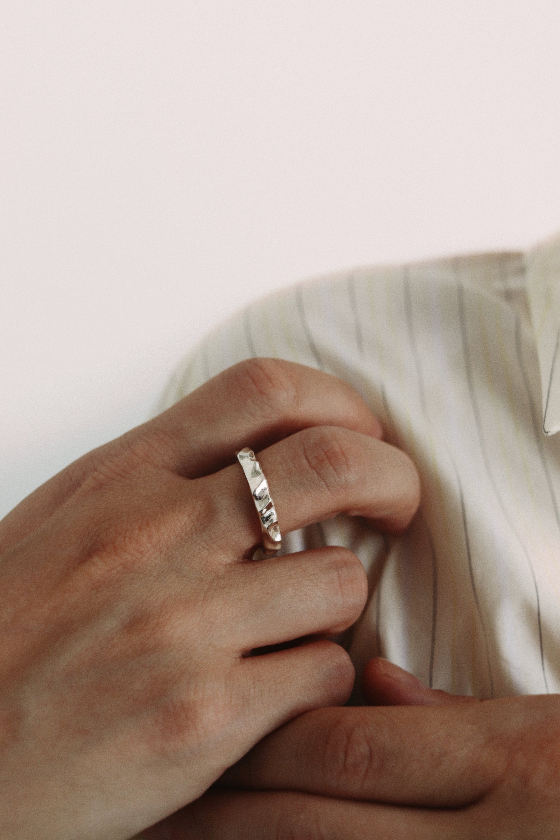 A close-up of a person's hand wearing a thin silver band ring with a textured, hammered or oxidized finish, resting near a striped cream shirt.
