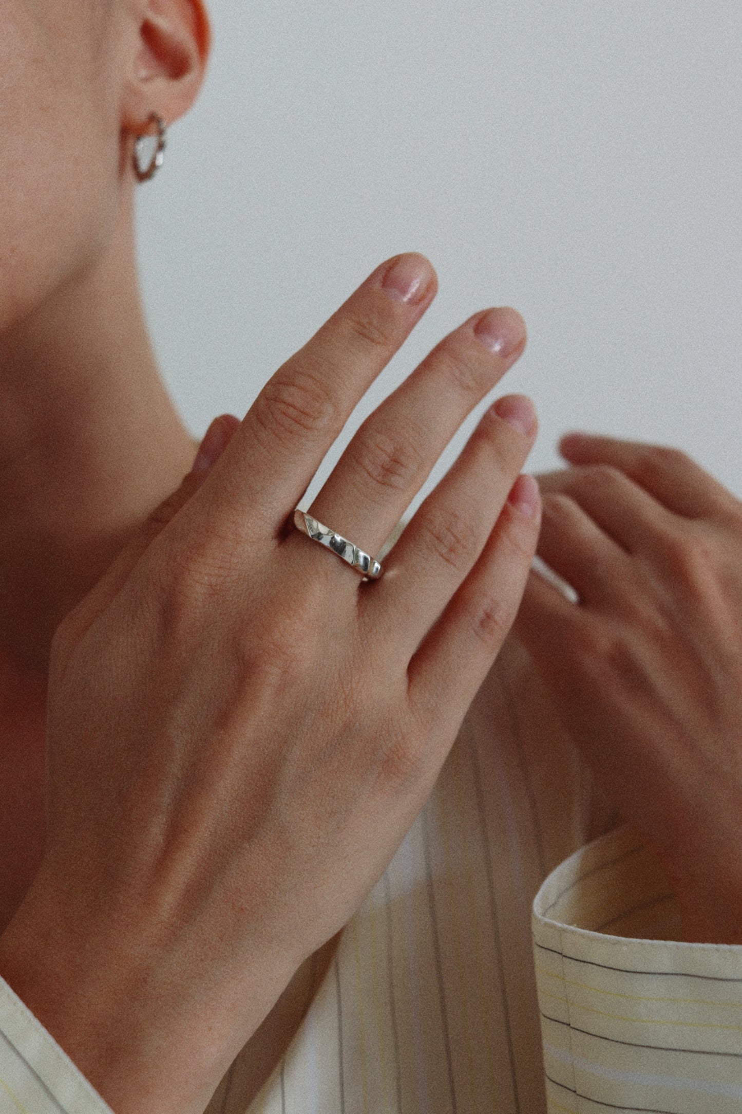 A close-up lifestyle shot of a person wearing a delicate silver band ring with small clear stones, shown on their ring finger. The person is wearing a cream-colored striped shirt, and the background is a soft, neutral light gray.