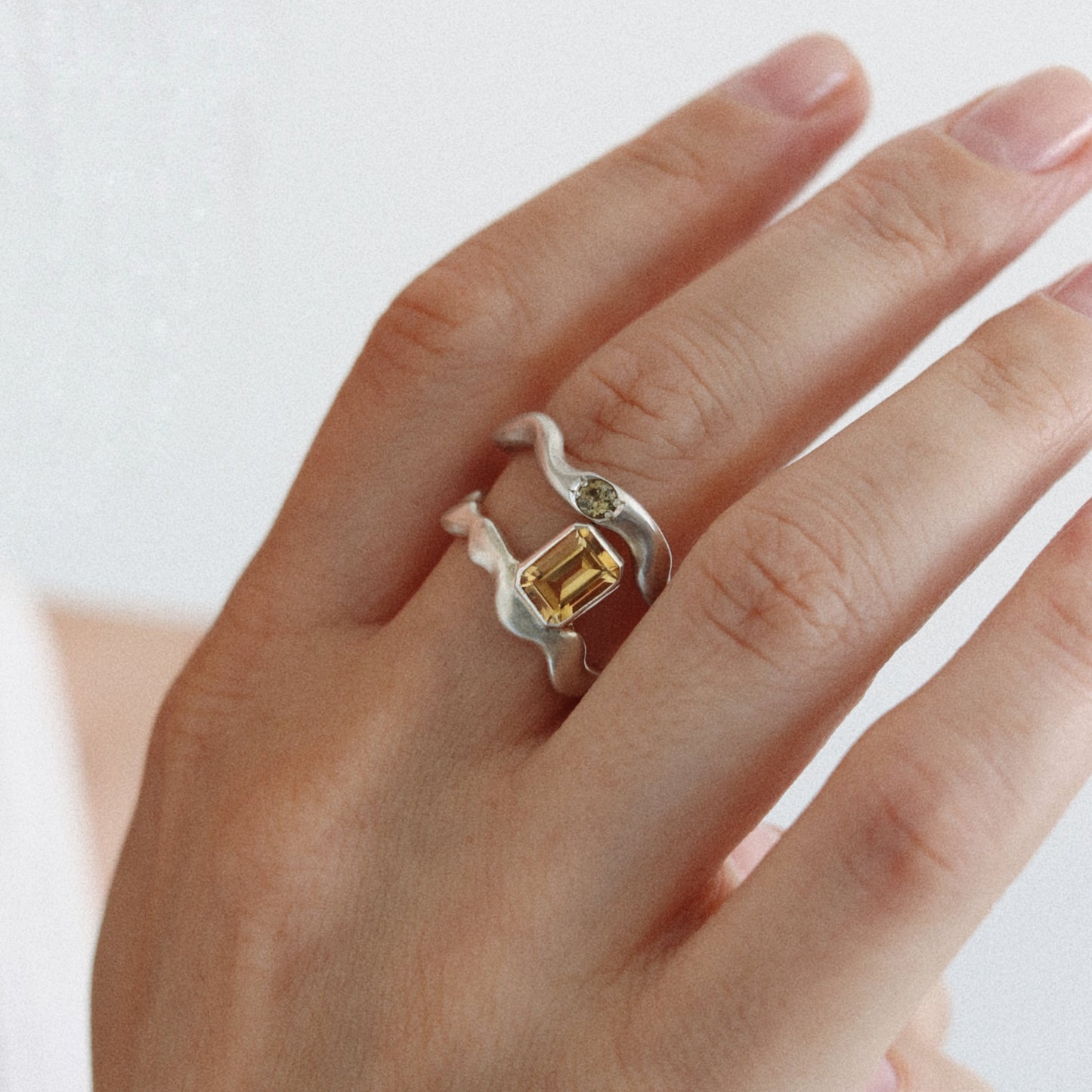 A close-up of a hand wearing a unique silver ring with a wavy band, a small green stone, and a central yellow rectangular gemstone.