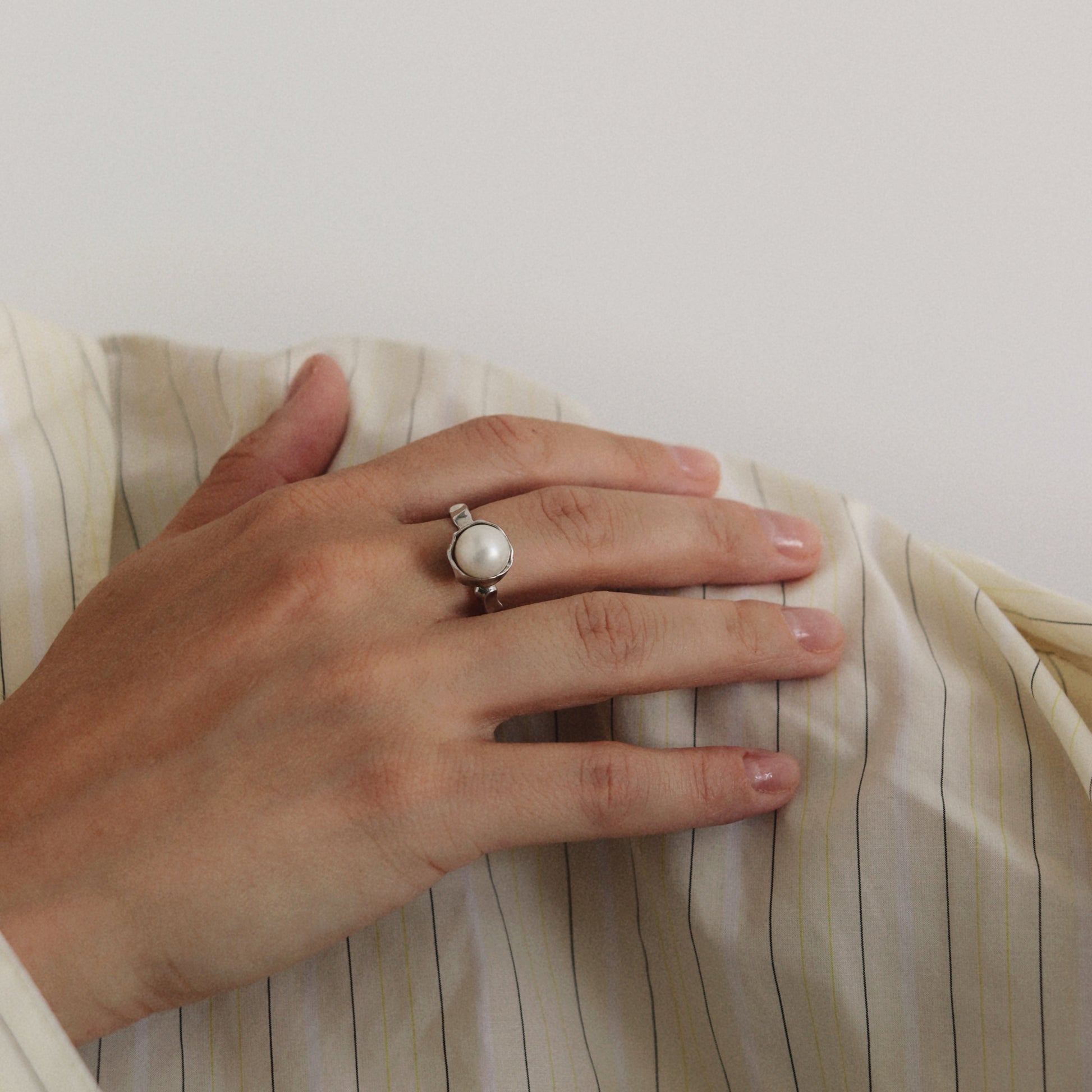 A silver ring with a large white pearl is worn on a person's ring finger, resting on a cream-colored pinstriped fabric against a plain light background.