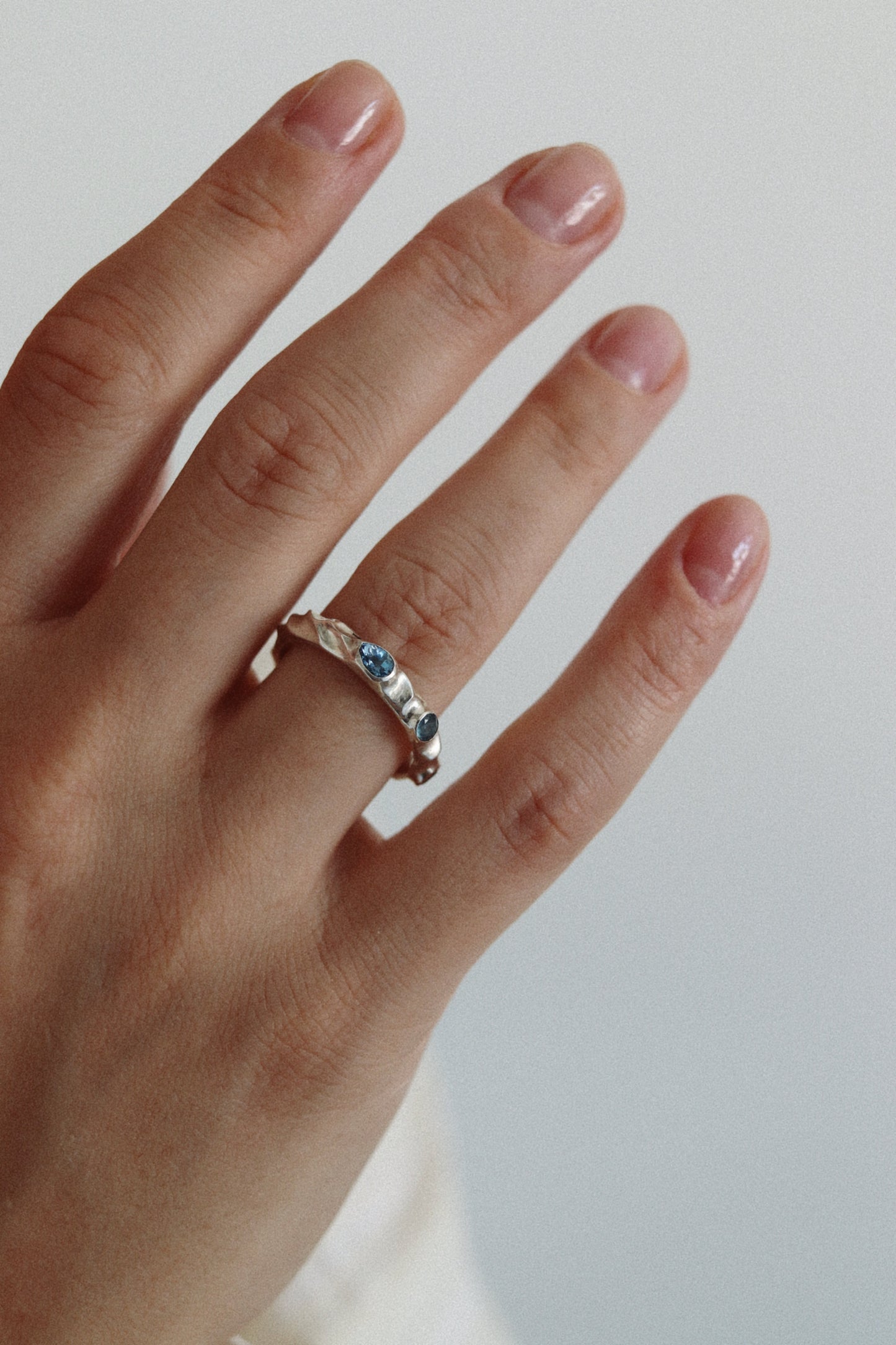 A close-up of a hand wearing a silver band ring with small blue gemstones, set against a plain light gray background.