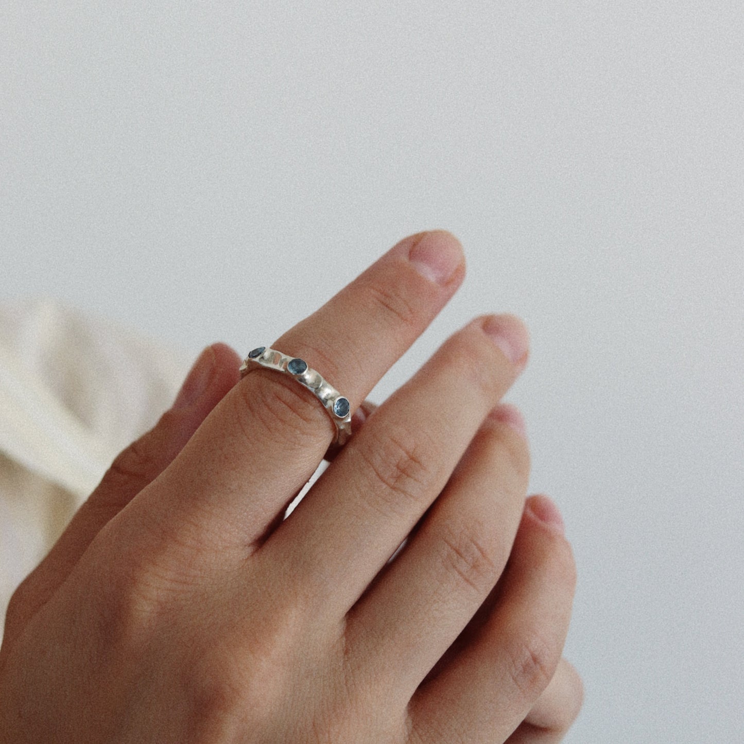 A close-up of a hand wearing a delicate silver band ring with small blue gemstones, set against a soft, light gray background.