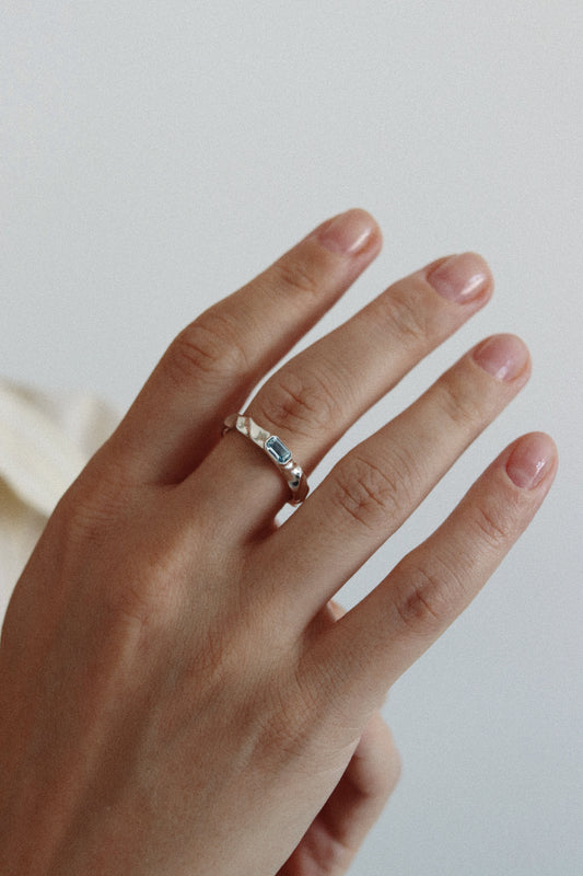 A close-up of a hand wearing a delicate silver band ring with a central blue stone, set against a soft, light gray background.