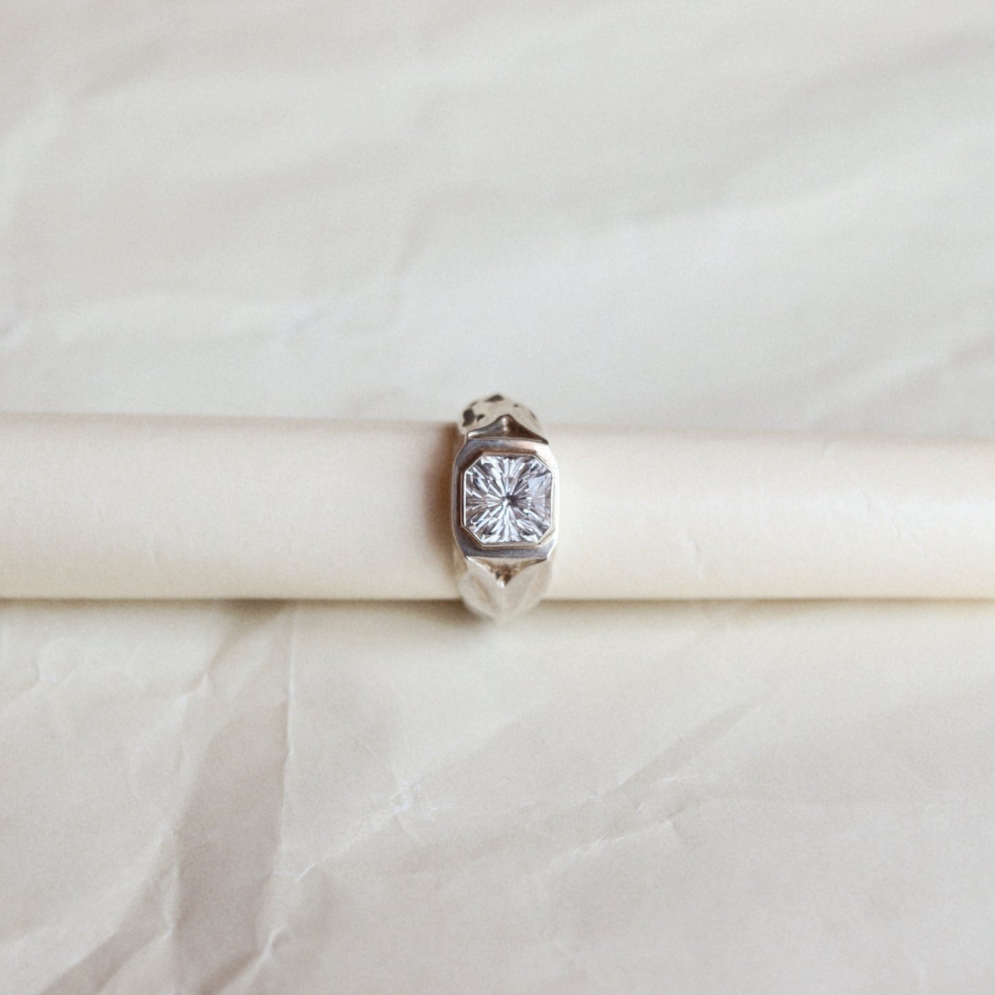 A silver ring with a square-cut clear gemstone, displayed on a cream-colored ring stand against a textured beige background.