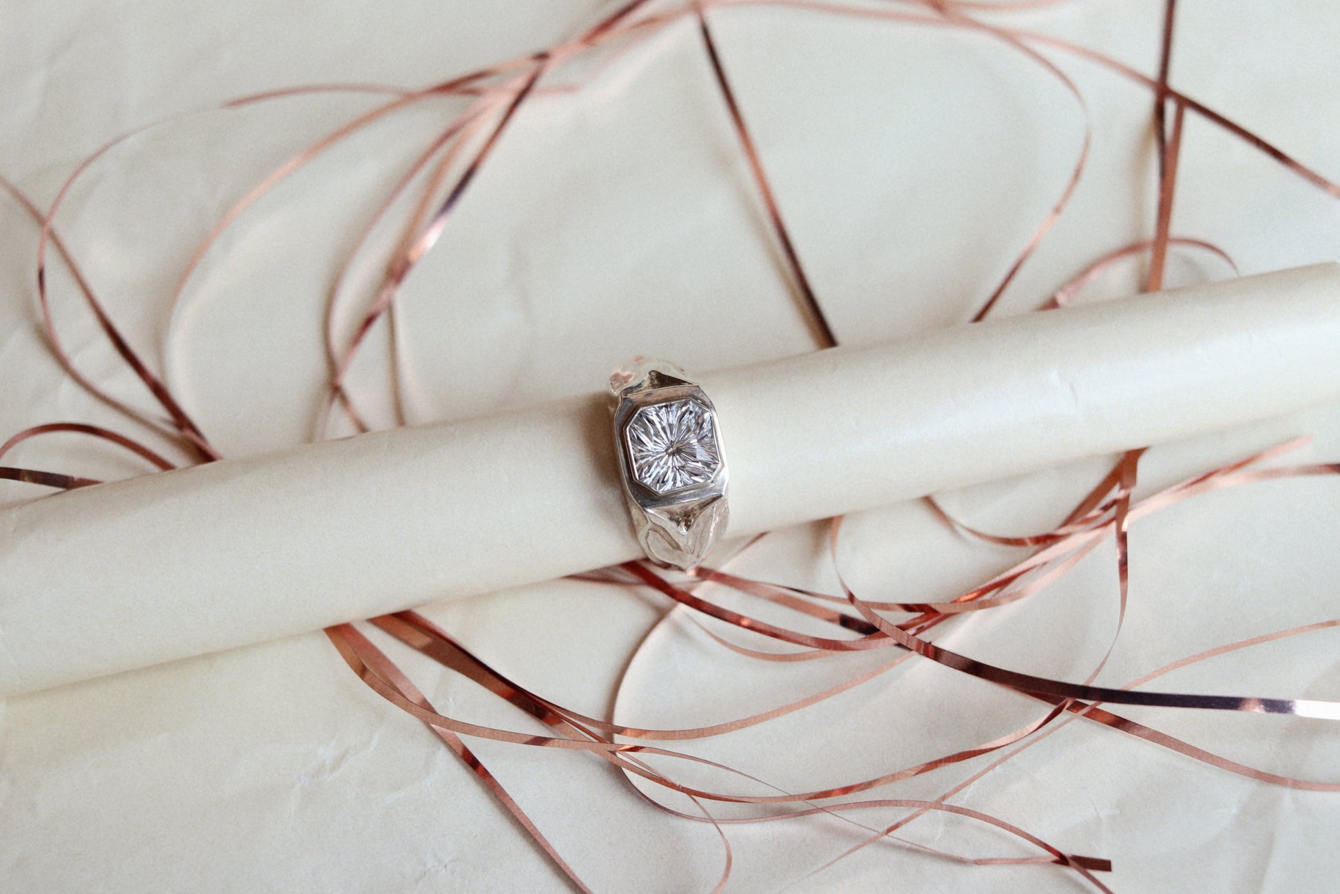 A silver-toned ring with a round clear gemstone, displayed on a cream-colored cylindrical stand with scattered rose gold ribbon.