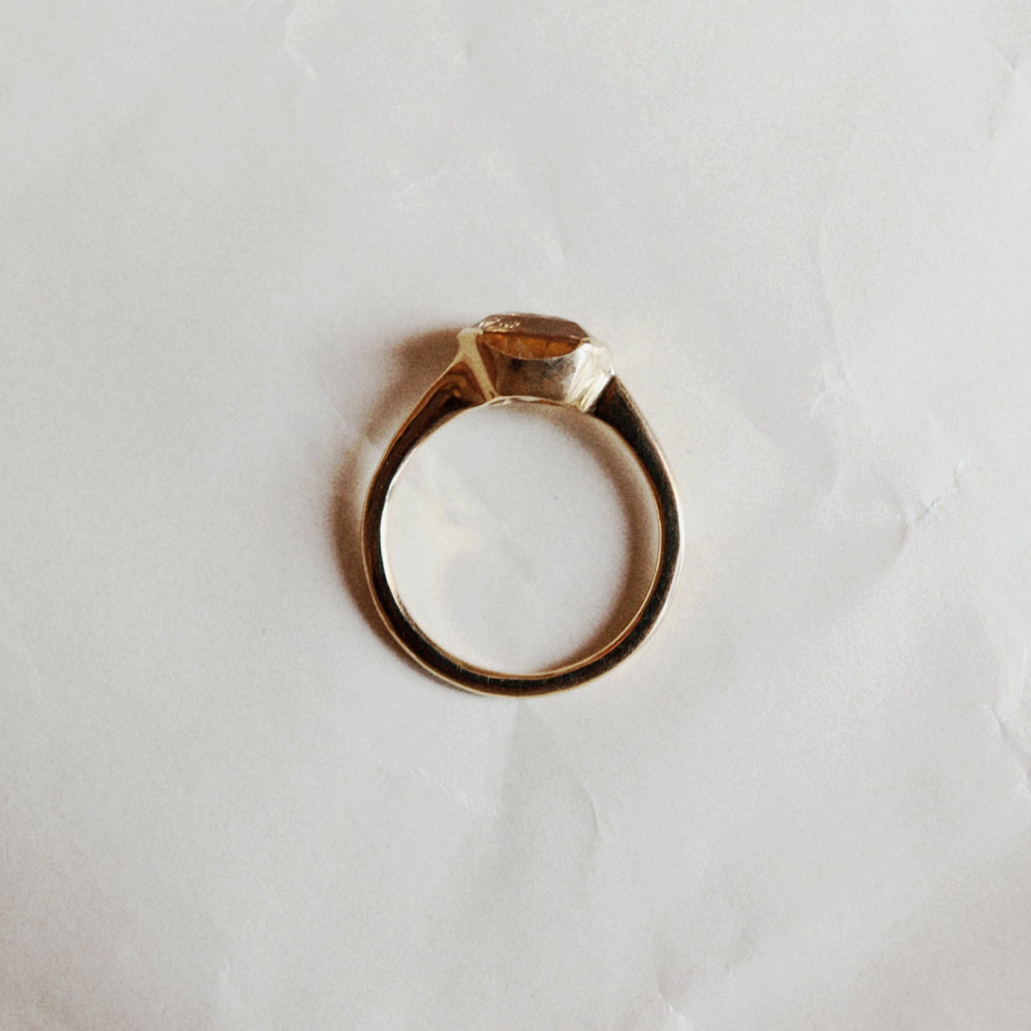 A gold ring with a small rectangular clear gemstone in a prong setting, photographed from above on a wrinkled white paper background.