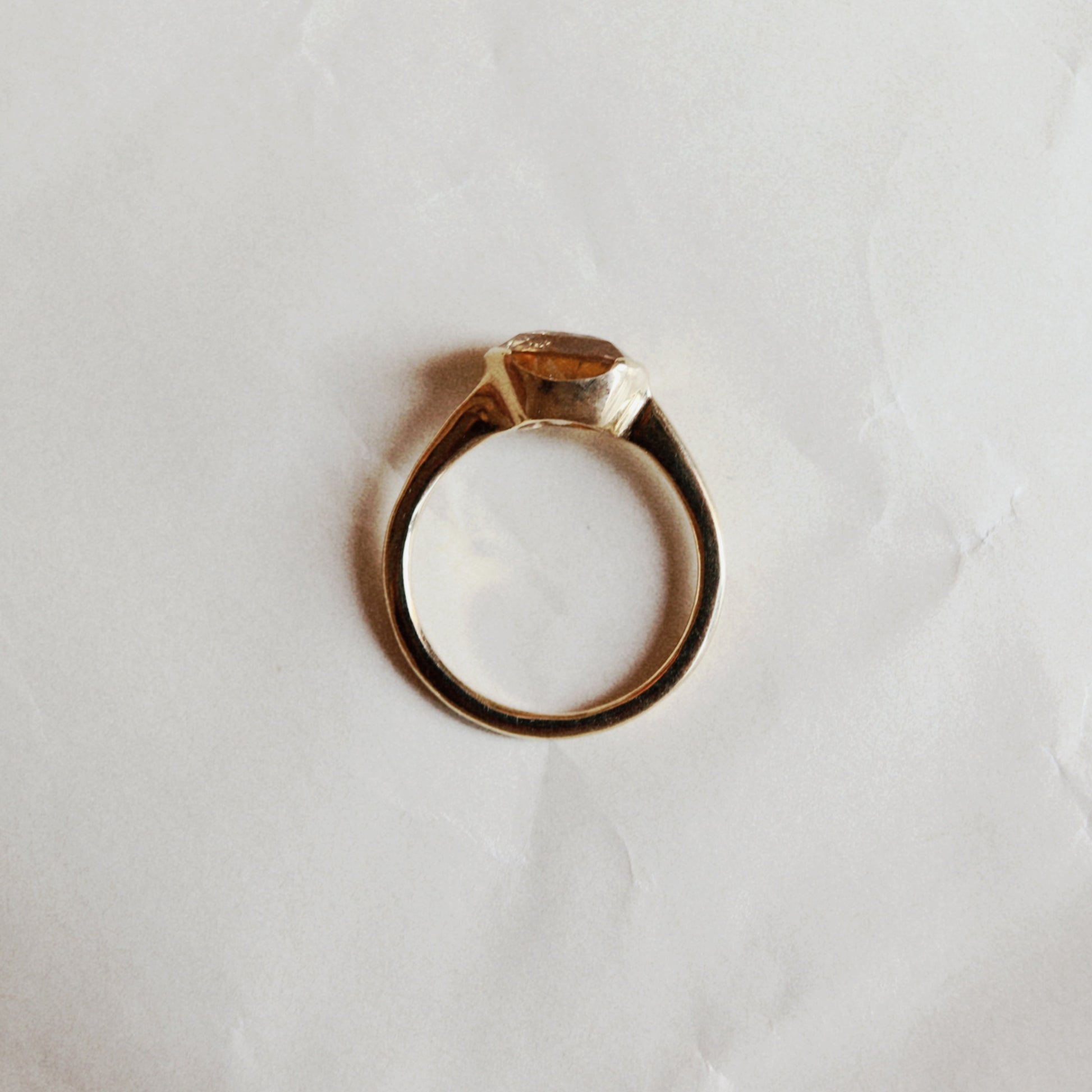 A gold ring with a small rectangular clear gemstone in a prong setting, photographed from above on a wrinkled white paper background.
