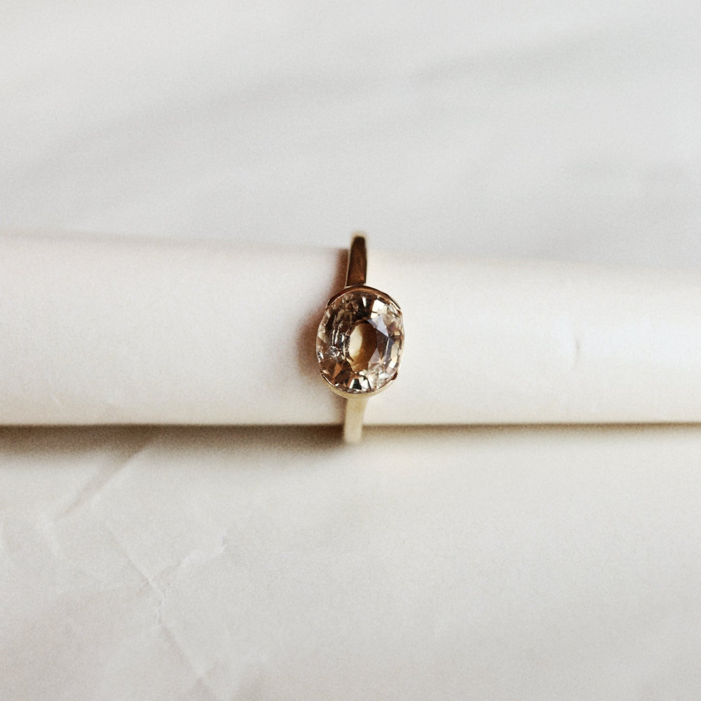 A gold ring with a small, square-cut clear gemstone in a prong setting, photographed from above on a textured, off-white paper background.