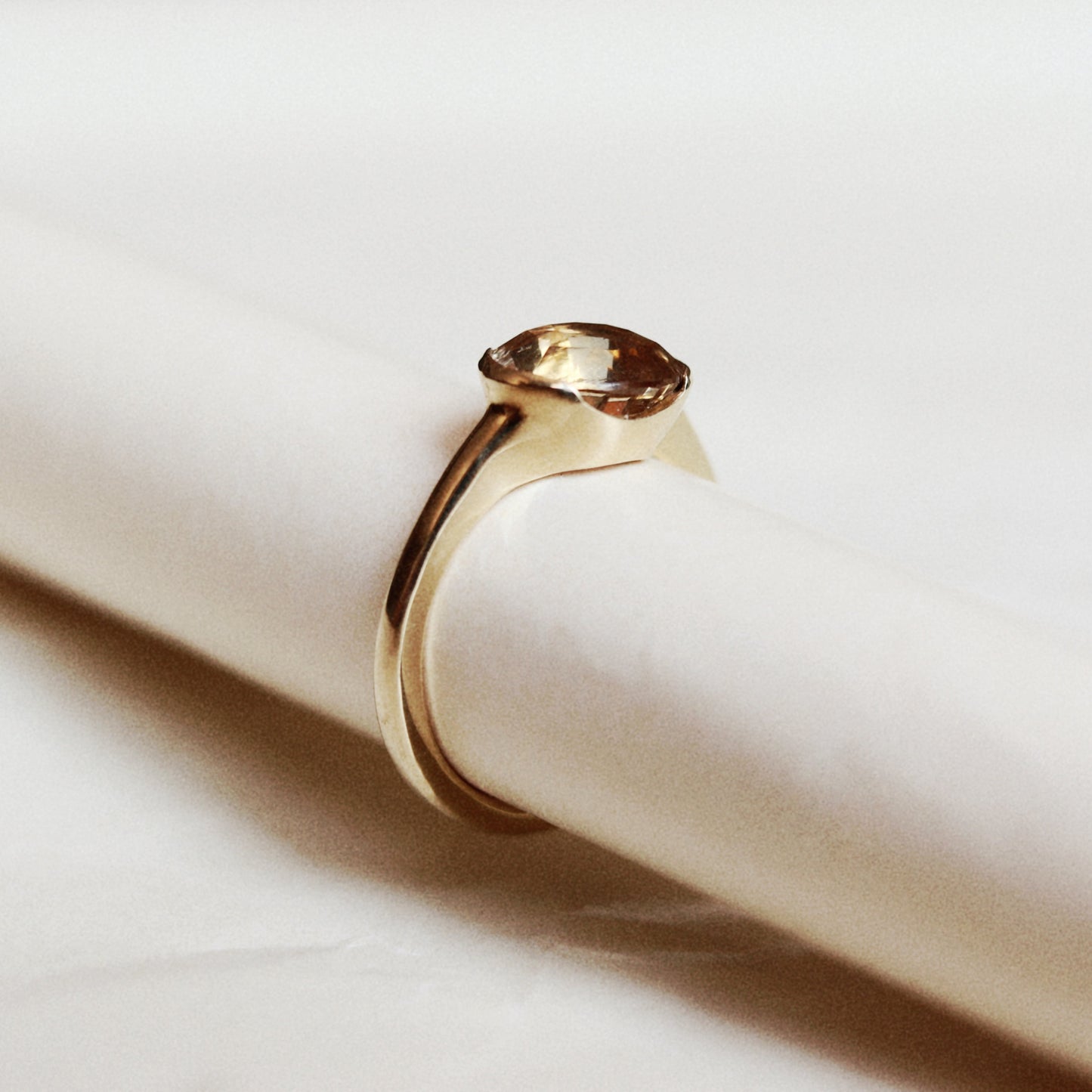 A gold ring with a marquise-cut brown gemstone, displayed on a white cylindrical ring stand against a soft white background.