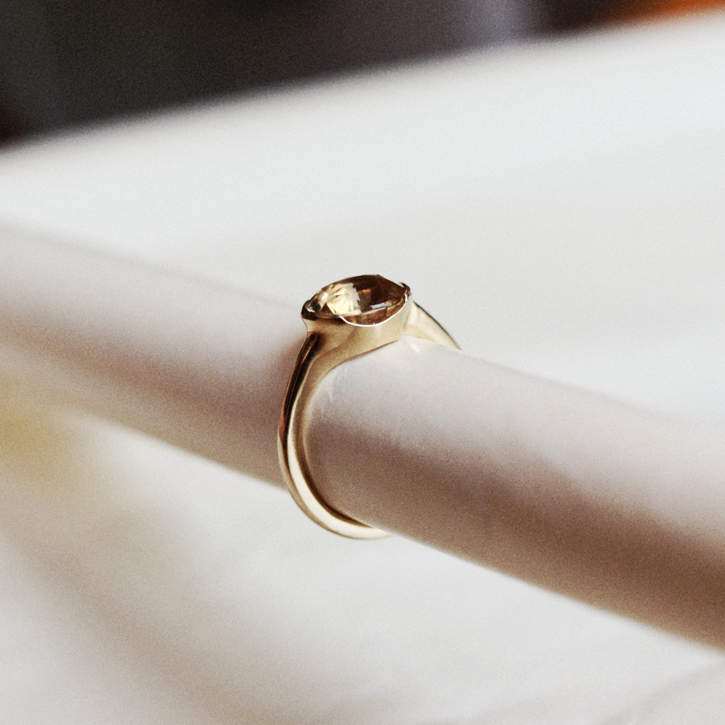 A gold ring with a bezel-set brown gemstone, displayed on a white cylindrical ring stand.